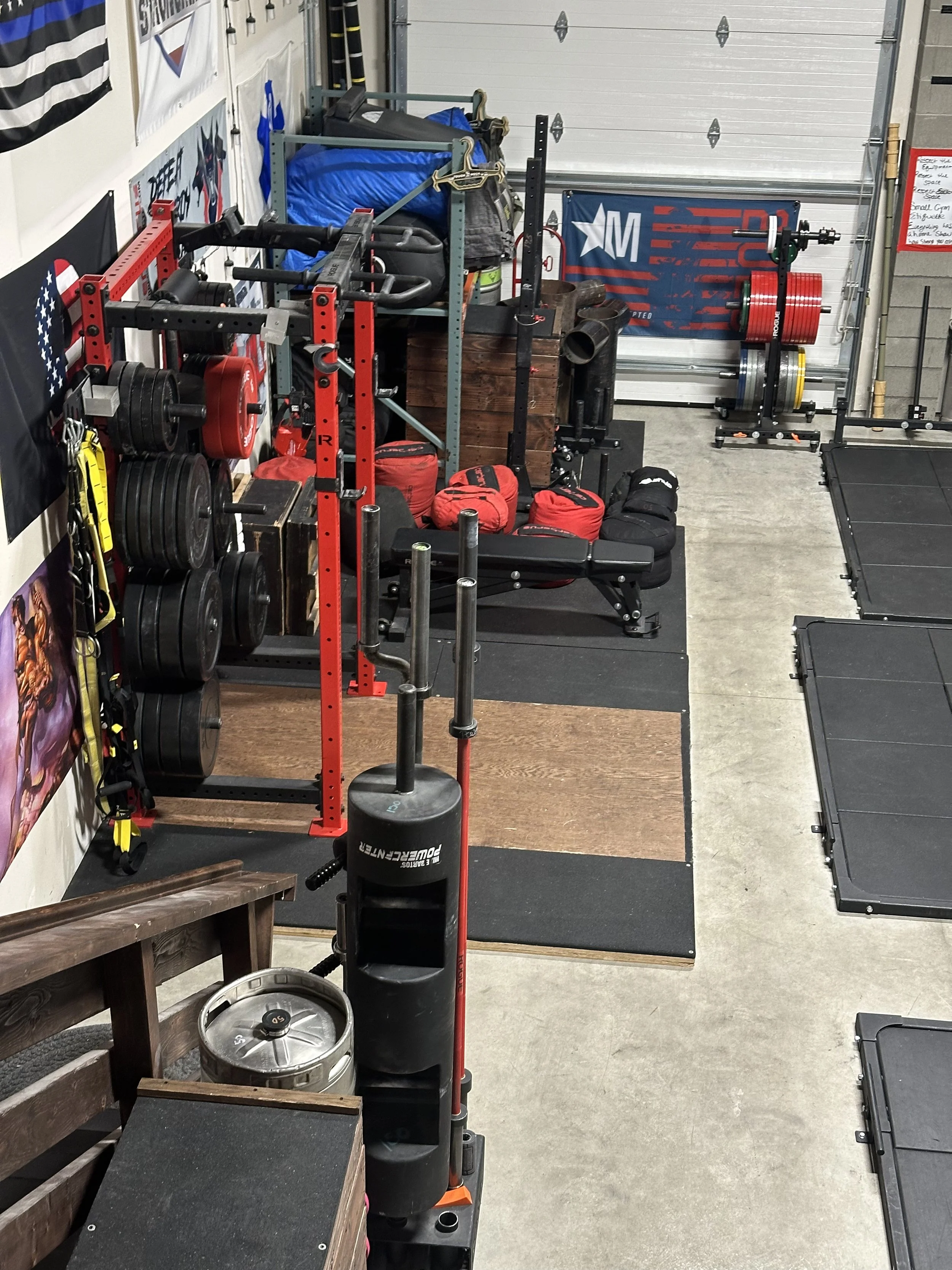 A workout space showcasing weightlifting equipment, including a red and black squat rack with weight plates, a bench, and a rack with red and yellow weight plates. There are black rubber mats on the floor, a white wall with posters, and a garage door