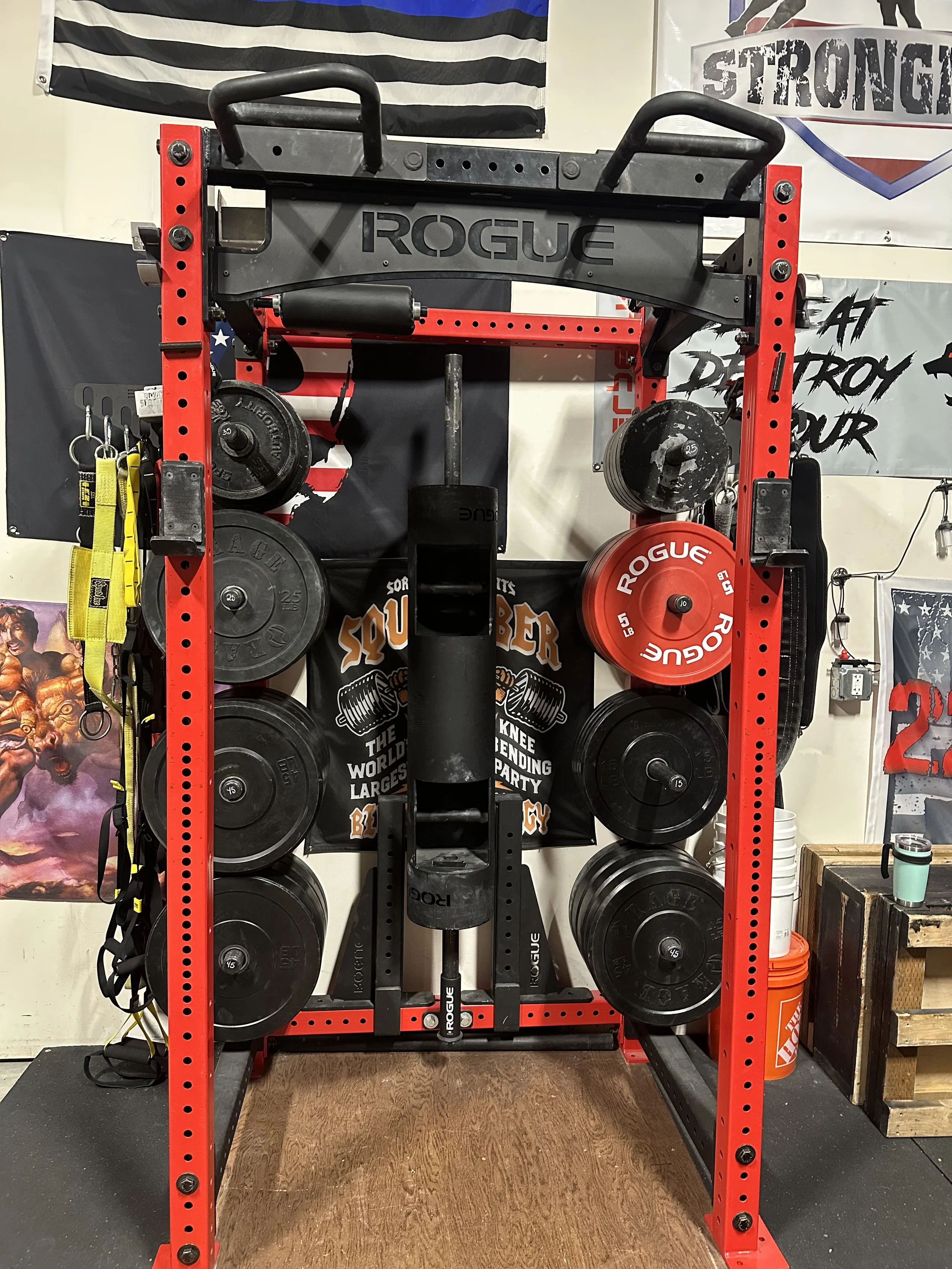 A red gym power rack with black and red weight plates, a black Rogue-branded pull-up bar, and various workout accessories in a gym setting.