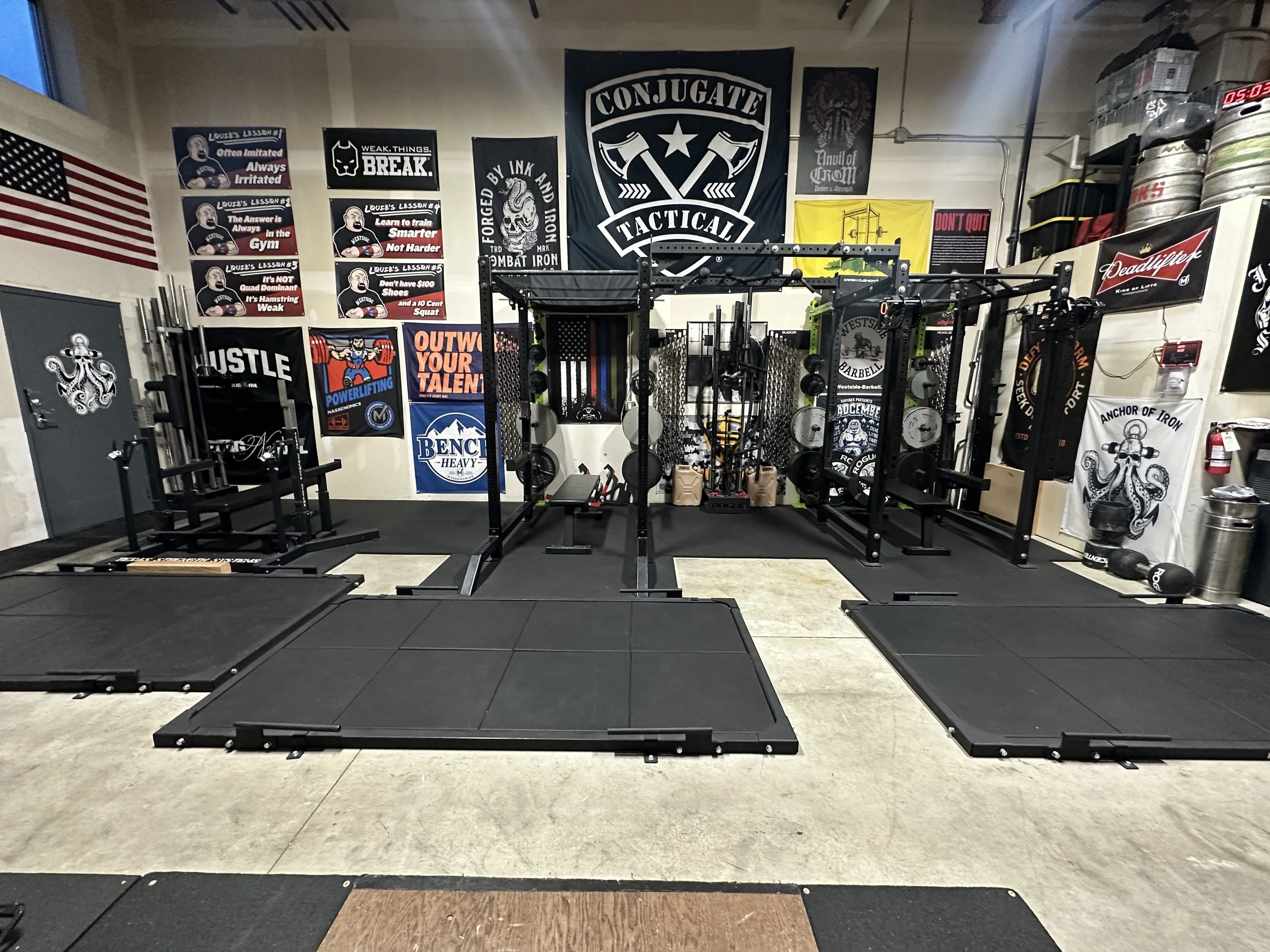A gym with various exercise equipment, patriotic flags, and motivational banners. The walls display black and white flags, and there are weights and squat racks in the center.
