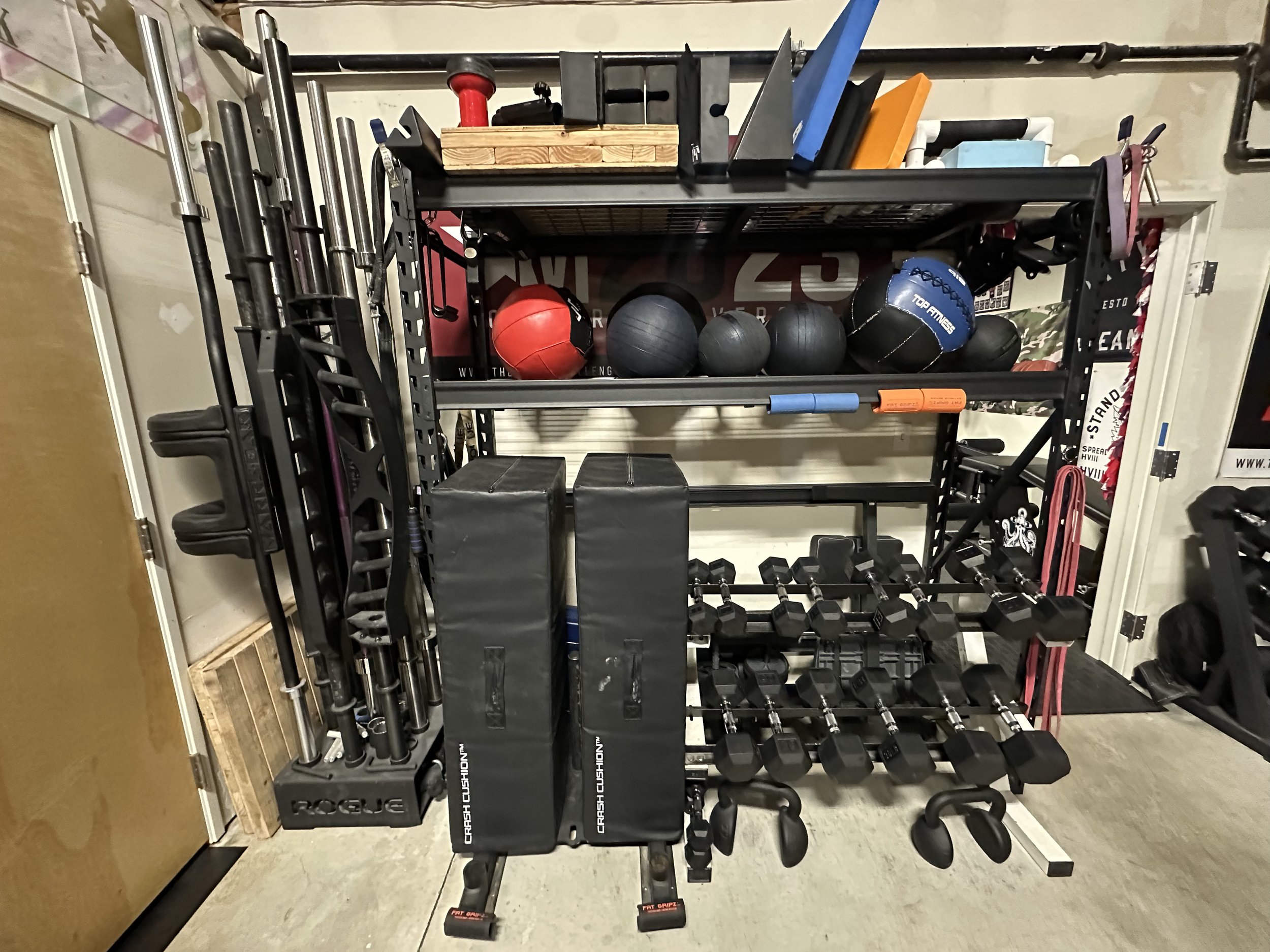A black metal workout rack holding various fitness equipment including barbells, medicine balls, kettlebells, foam rollers, and resistance bands in a home gym.