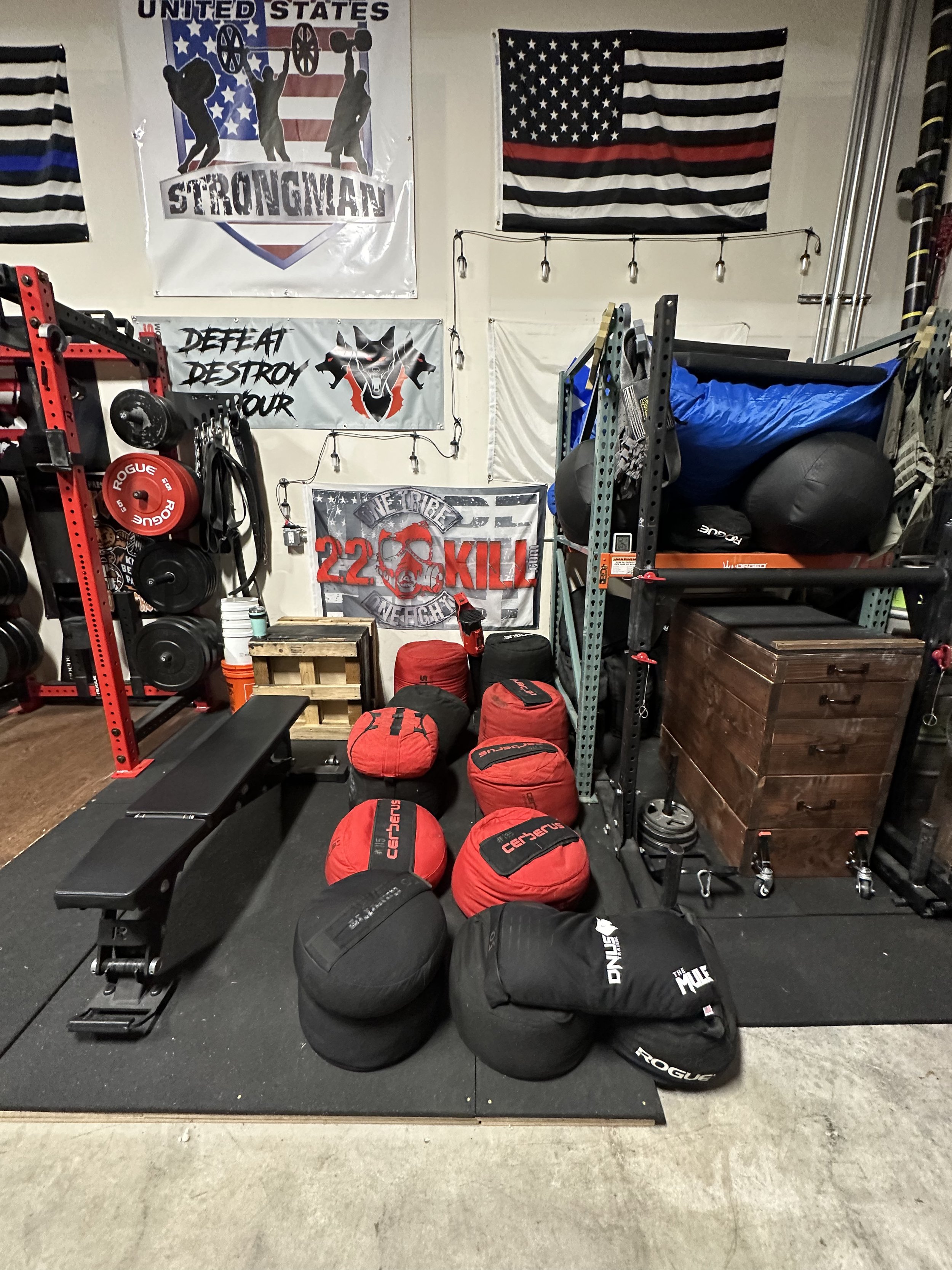 A gym corner with flags and banners, including a U.S. flag with a black and red stripe, a Strongman event banner, and a 'Defeat Destroy Your' banner. Fitness equipment such as weights, kettlebells, medicine balls, and a weight bench are arranged on t