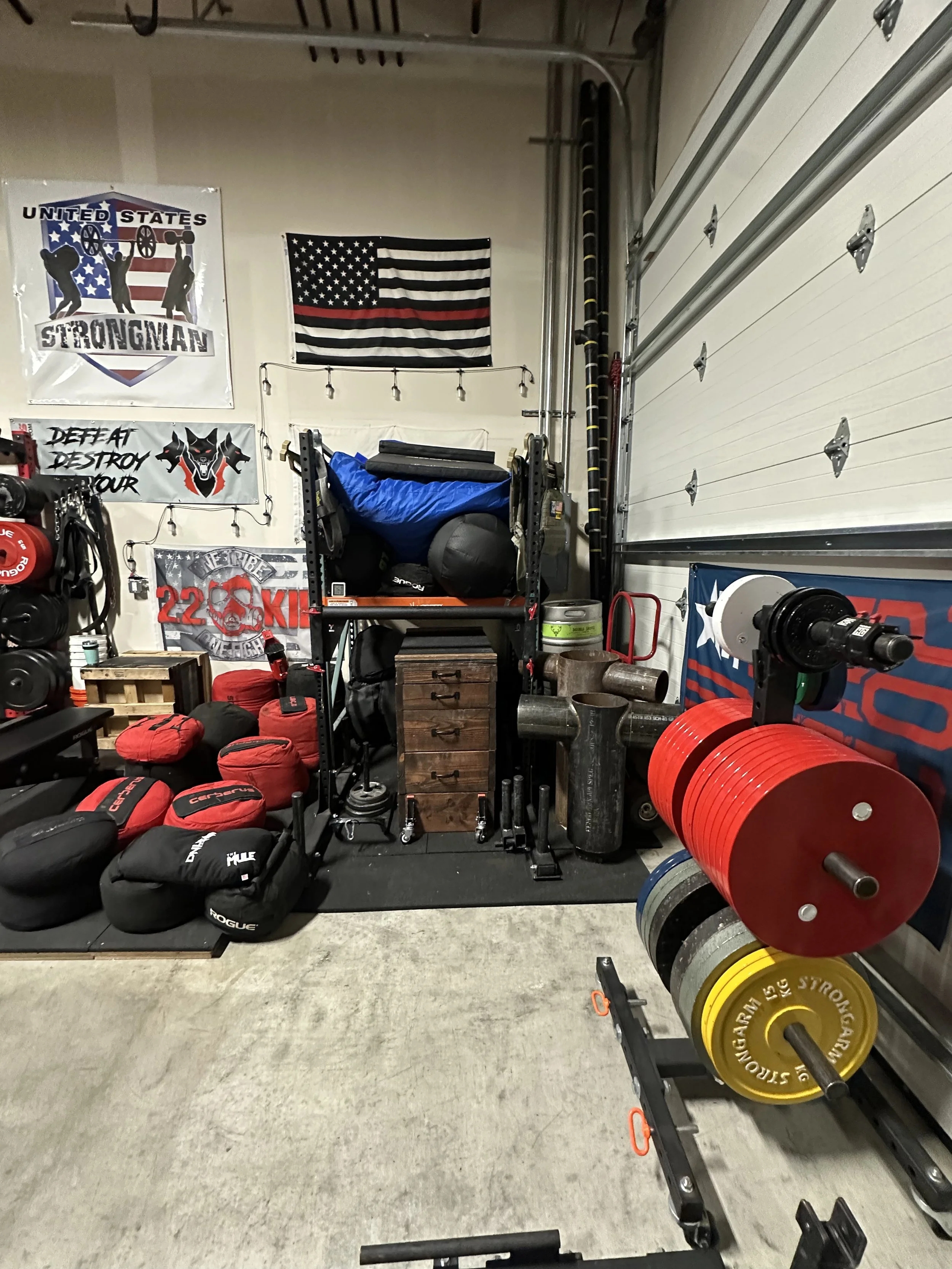 A home gym with weightlifting equipment, including barbells, weight plates, and sandbags, with motivational posters and flags on the walls.