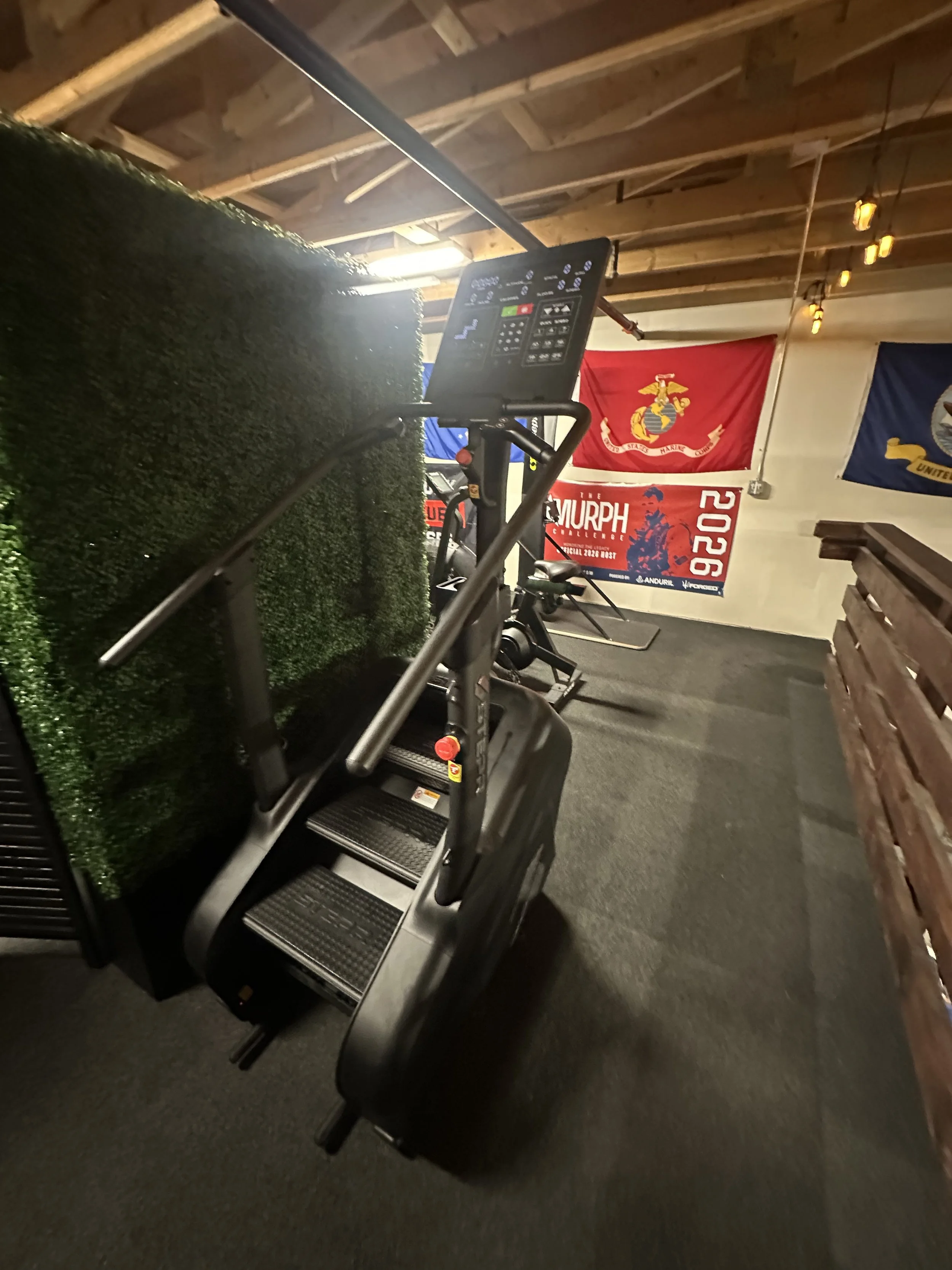 Gym with stair climber machine, tennis court barrier, and flags of the United States Marine Corps and the United States Navy on the wall.