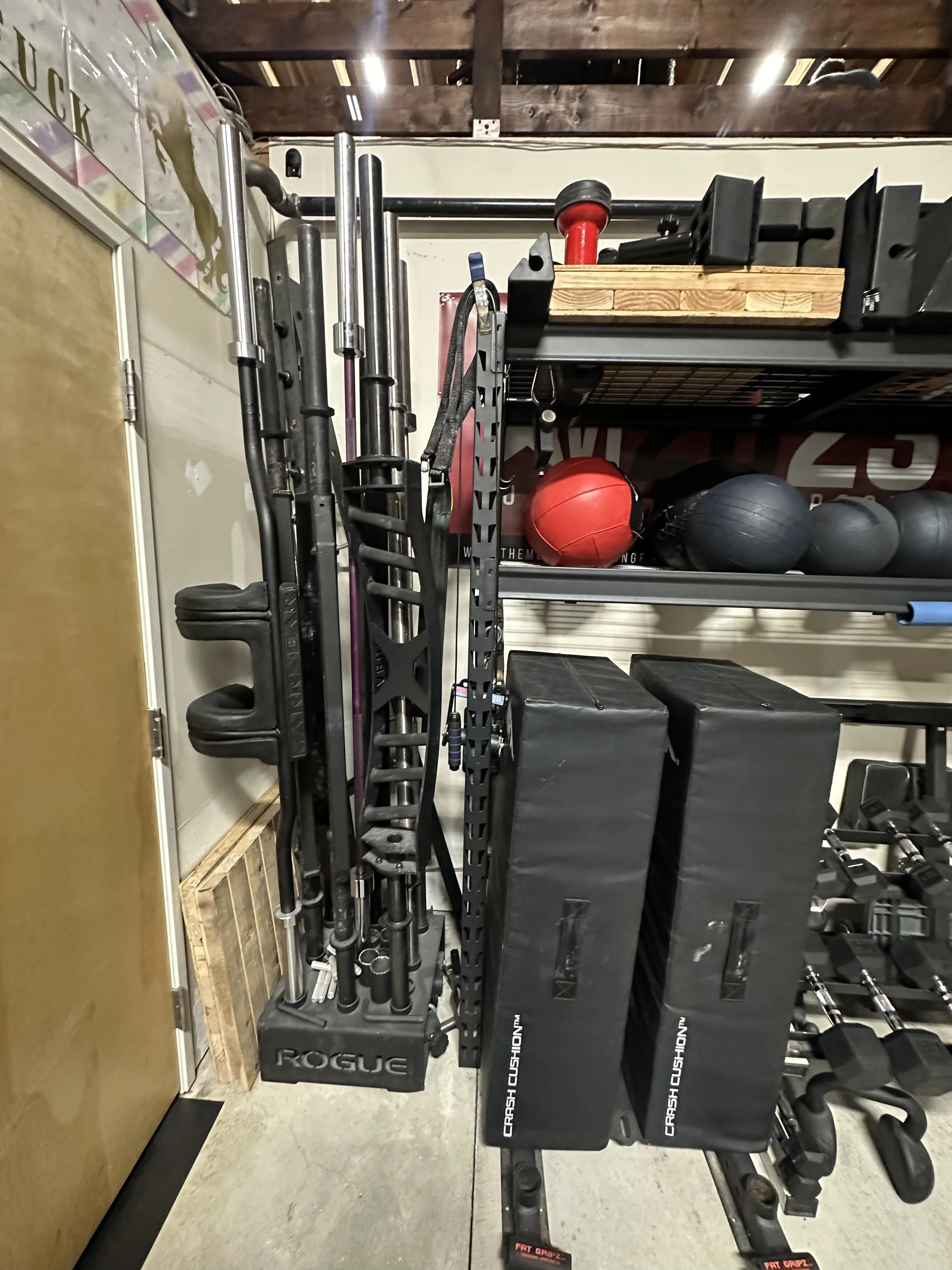 A storage area with various fitness equipment, including black and red medicine balls, black storage boxes labeled 'CRASH CUSHION WM,' barbells, and weight plates, along with a rack of long metal bars and accessories.
