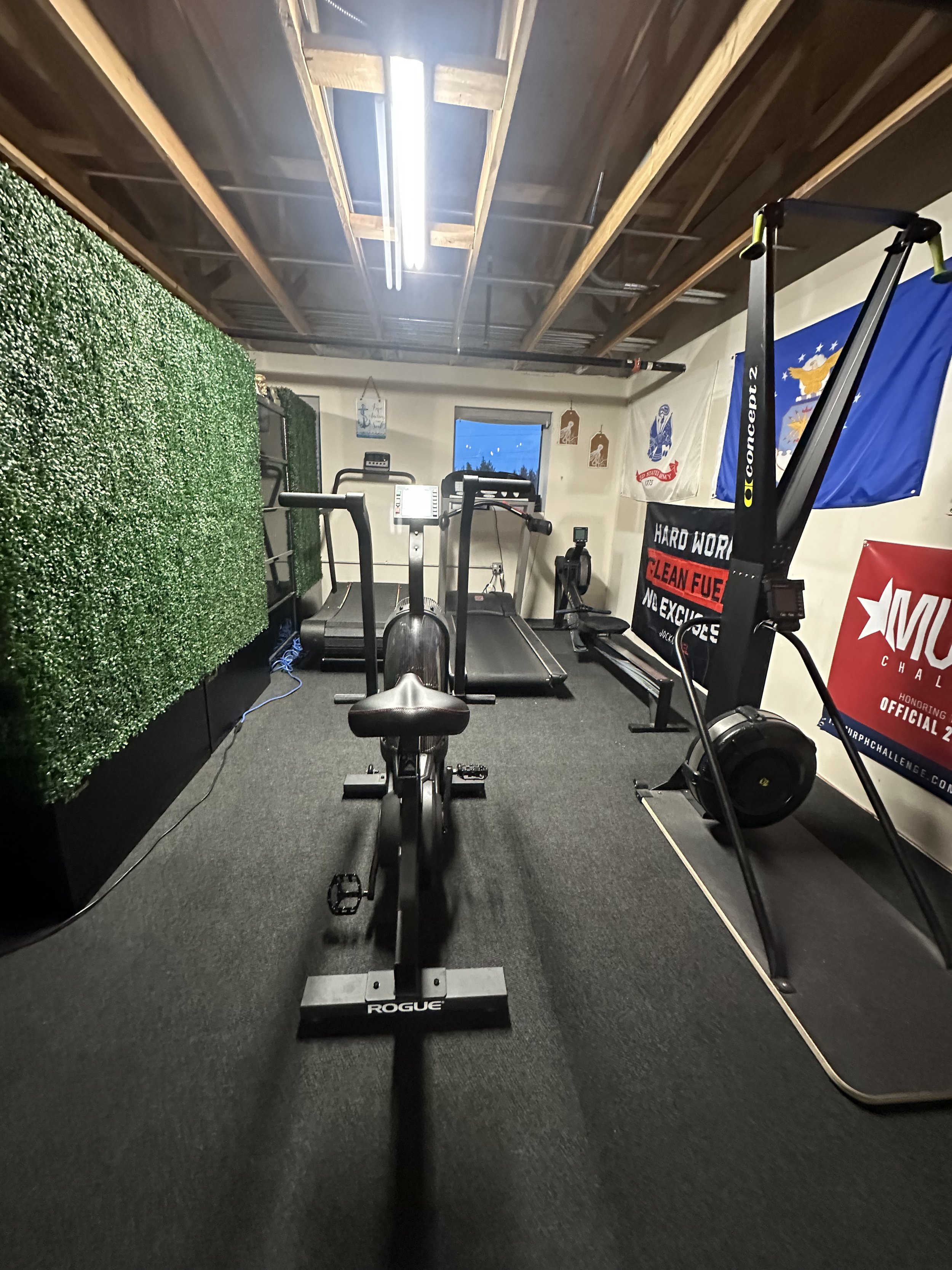 Home gym with a treadmill, exercise bike, and a rowing machine, with flags and motivational banners on the walls, and a green wall divider on the left side.