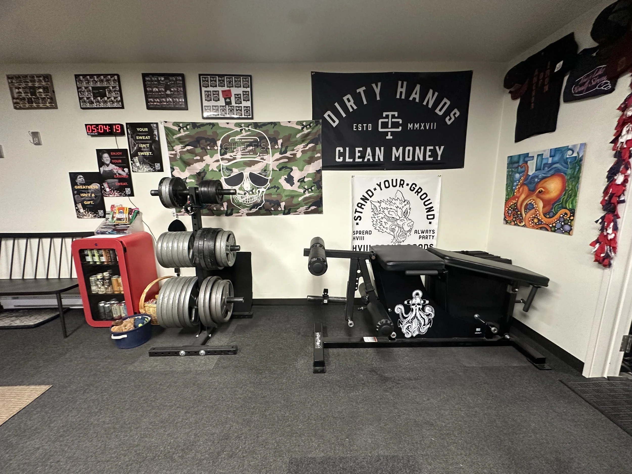 A home gym with weightlifting equipment including a barbell with weights on a rack, a treadmill, and various wall art. The wall displays several posters, a large skull flag with camouflage background, and other flags and artwork. There is a digital c