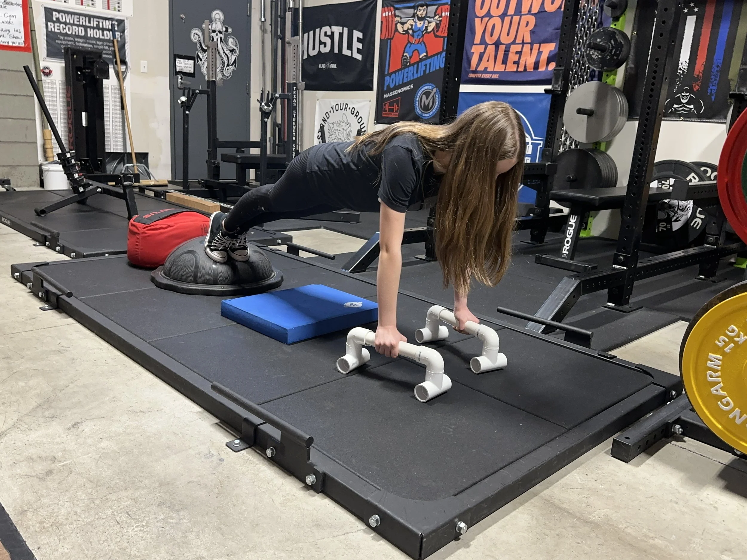 A woman in a gym performing a push-up with her hands on PVC pipes on a rubber mat. There are weightlifting equipment, plates, and motivational posters on the wall behind her.