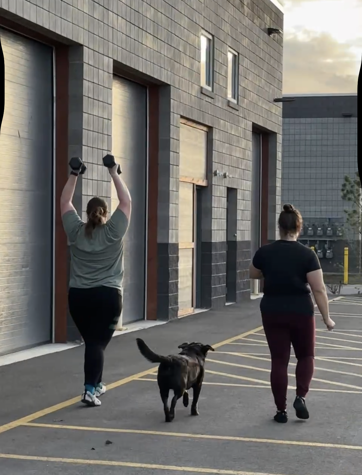 Two women and a black dog walking outside a commercial building during sunset. One woman is lifting dumbbells, the other is walking nearby, and the dog is walking between them.