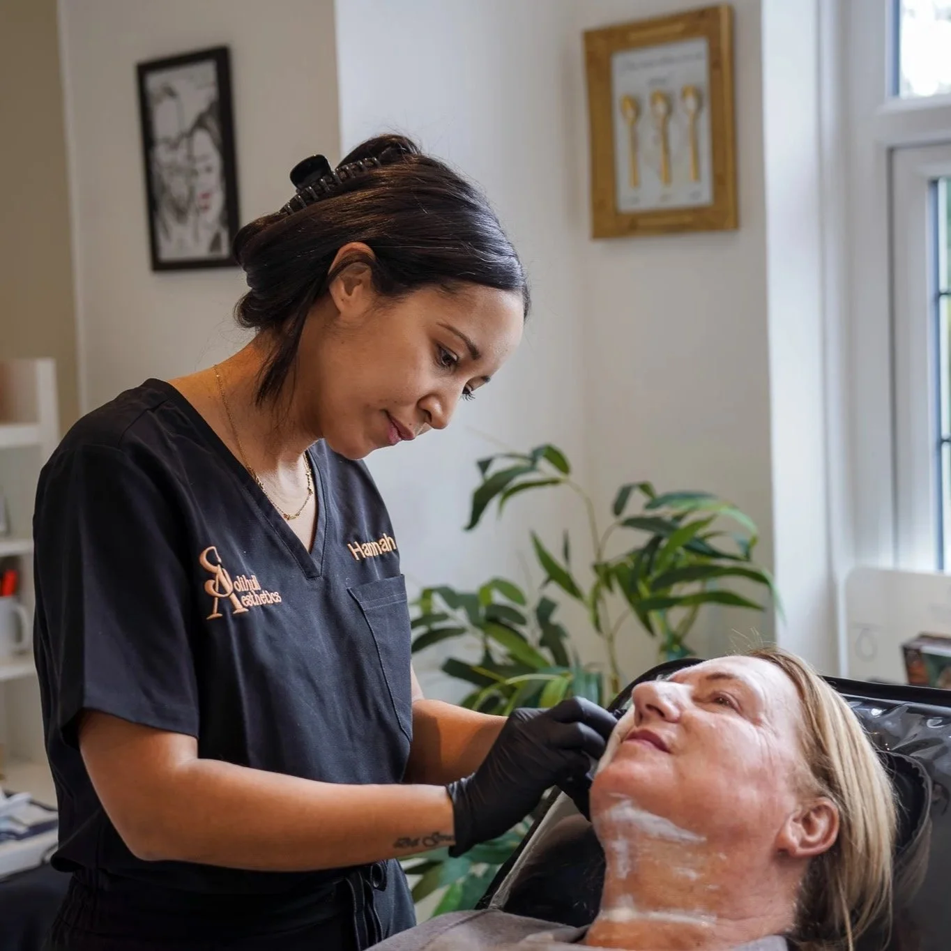 A skincare professional performing a facial on a woman lying back with a white cream on her face in a spa or clinic setting.