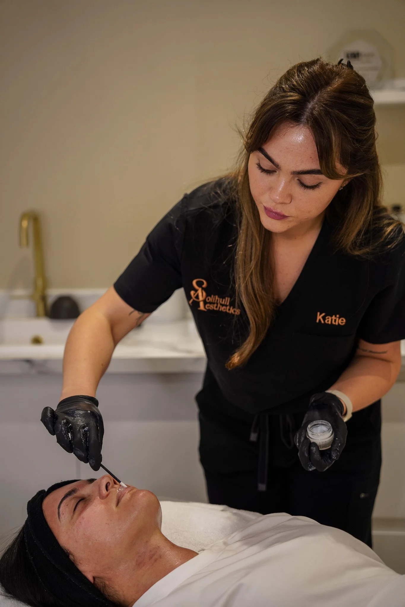 A woman receiving a facial treatment from a professional in a spa or clinic setting. The professional is wearing black gloves and appears to be applying a product or treatment near the woman's face, who is lying down with her eyes closed.