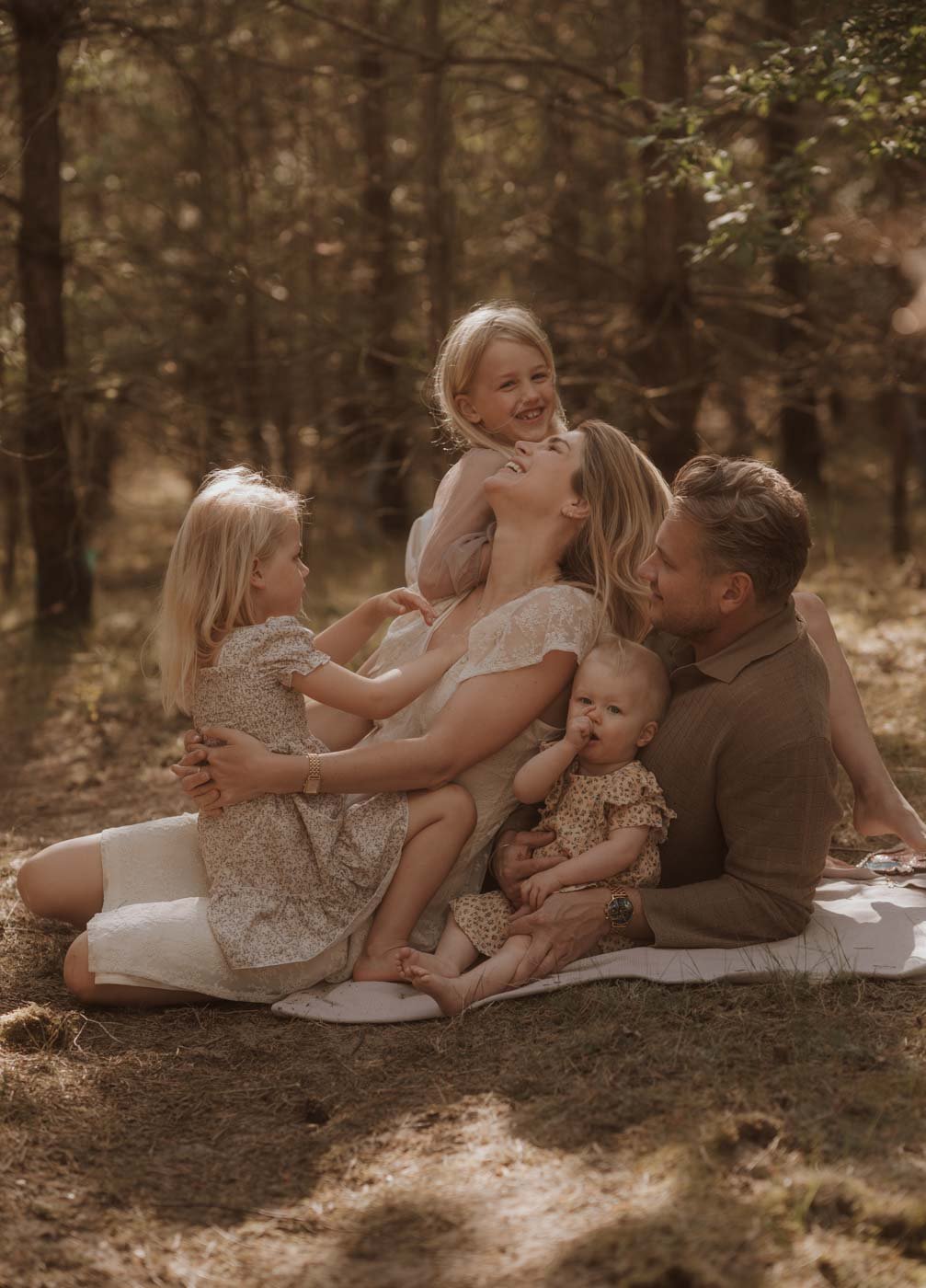Een gezin met drie kinderen lacht samen tijdens een lente fotoshoot in het bos van Soestduinen, vastgelegd door Ara Schrijvers, gezinsfotograaf in Utrecht.