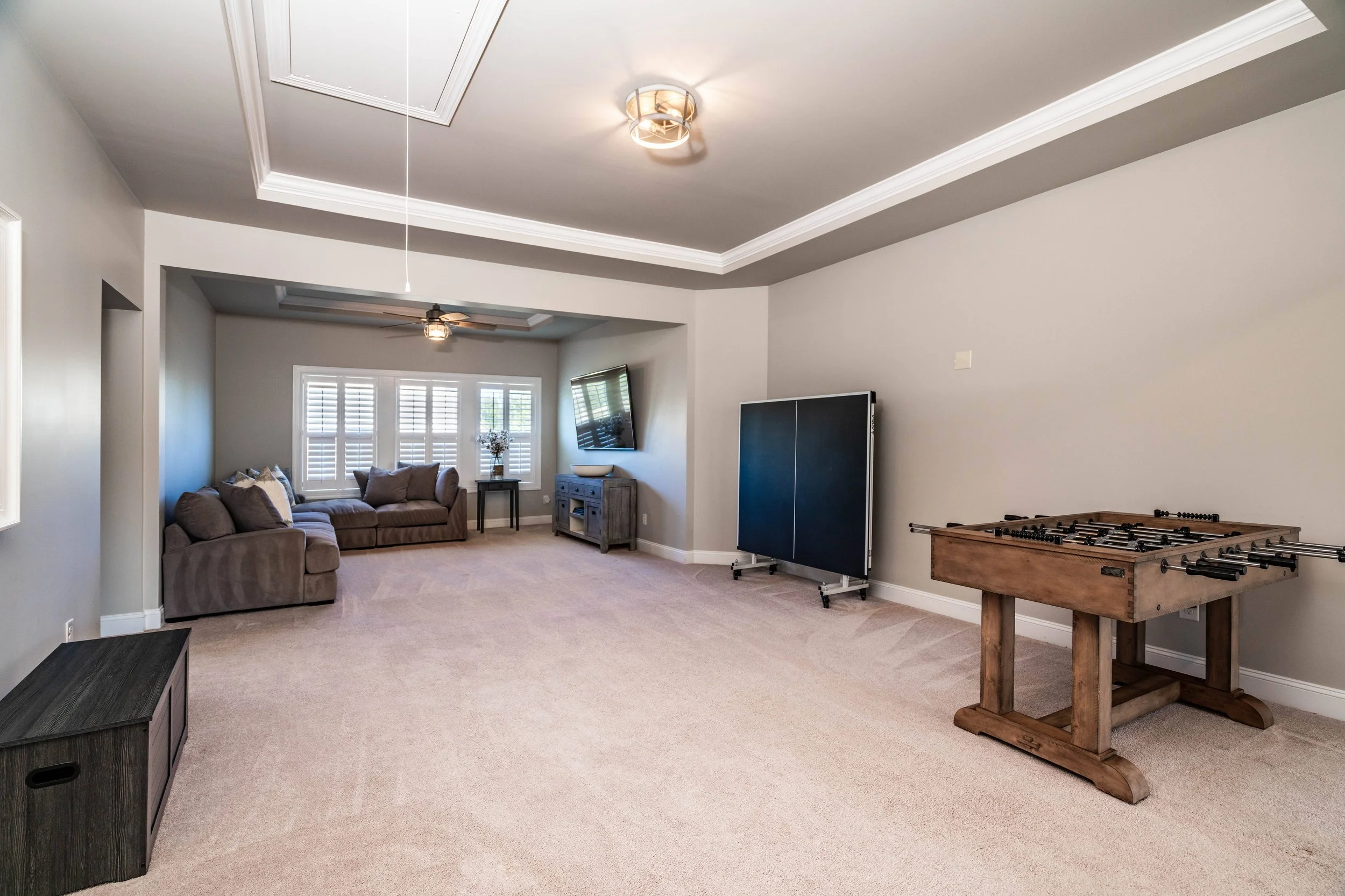 A spacious, carpeted game room with beige walls features a foosball table and folded ping pong table. A cozy seating area with sofas is under a ceiling fan.