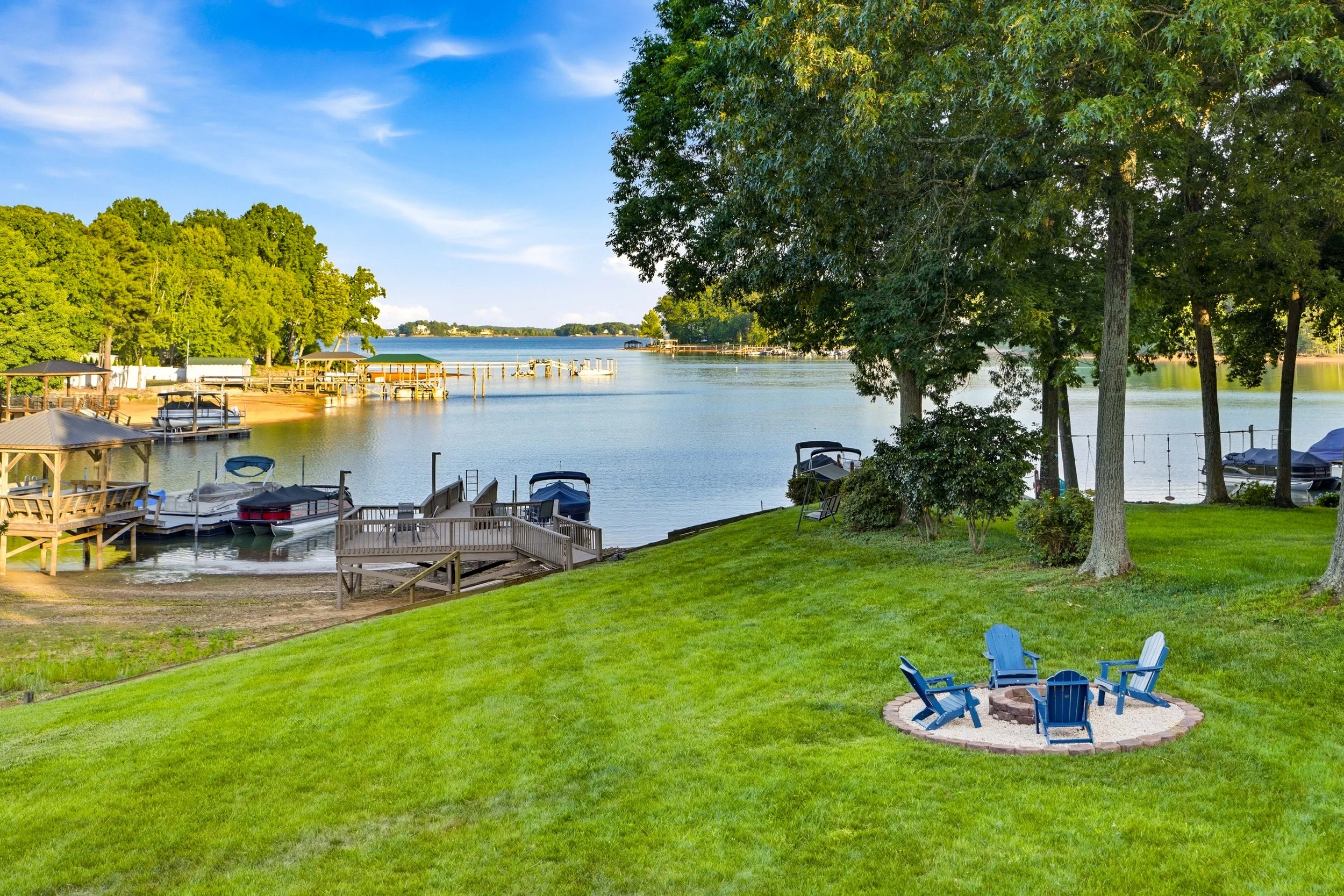 A grassy lawn with four blue Adirondack chairs around a stone fire pit, overlooking a lake with boat docks and trees under a bright blue sky.