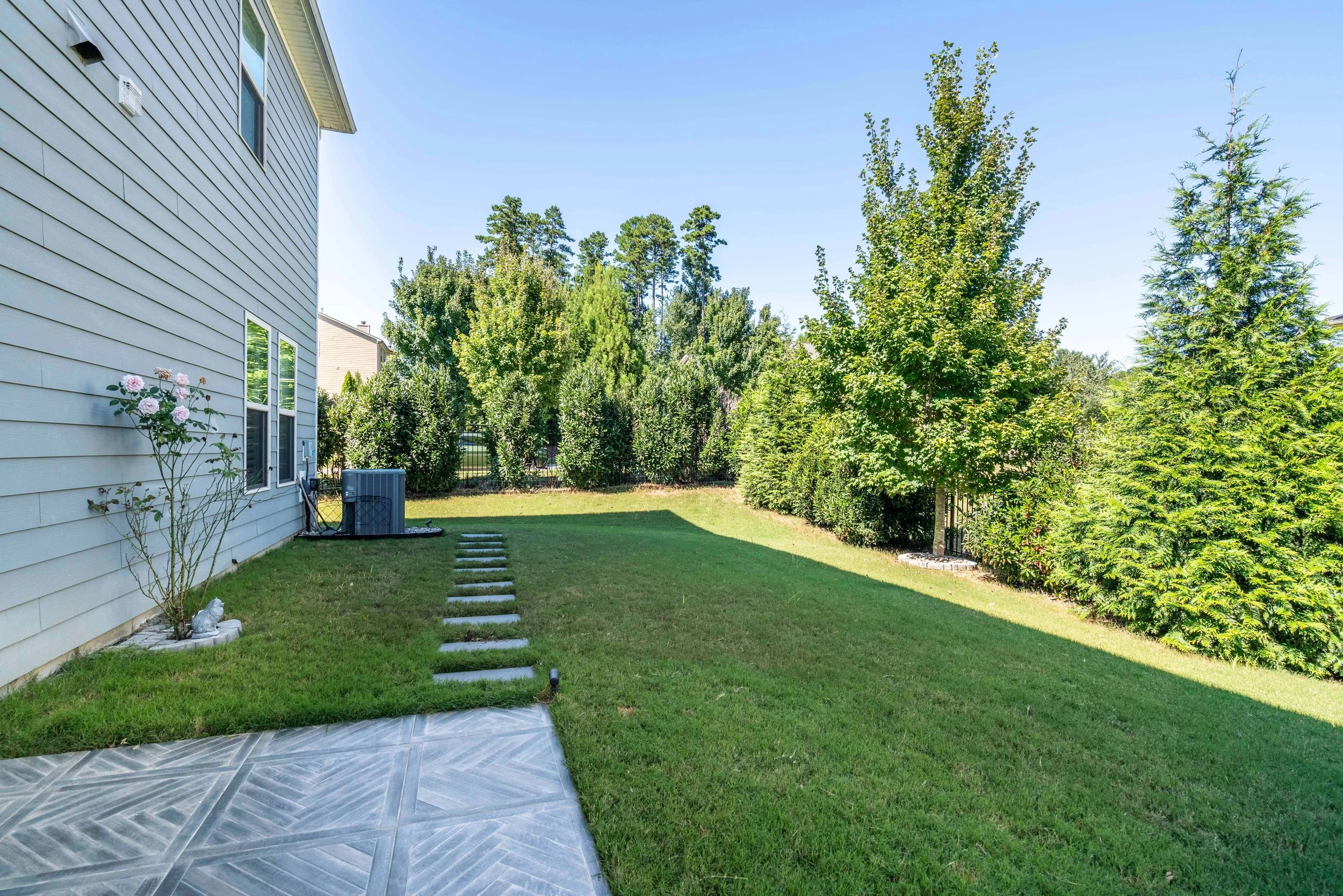Backyard with green grass, bordered by tall green trees. A light-colored house on the left. Stone path leads to a distant air conditioner unit.