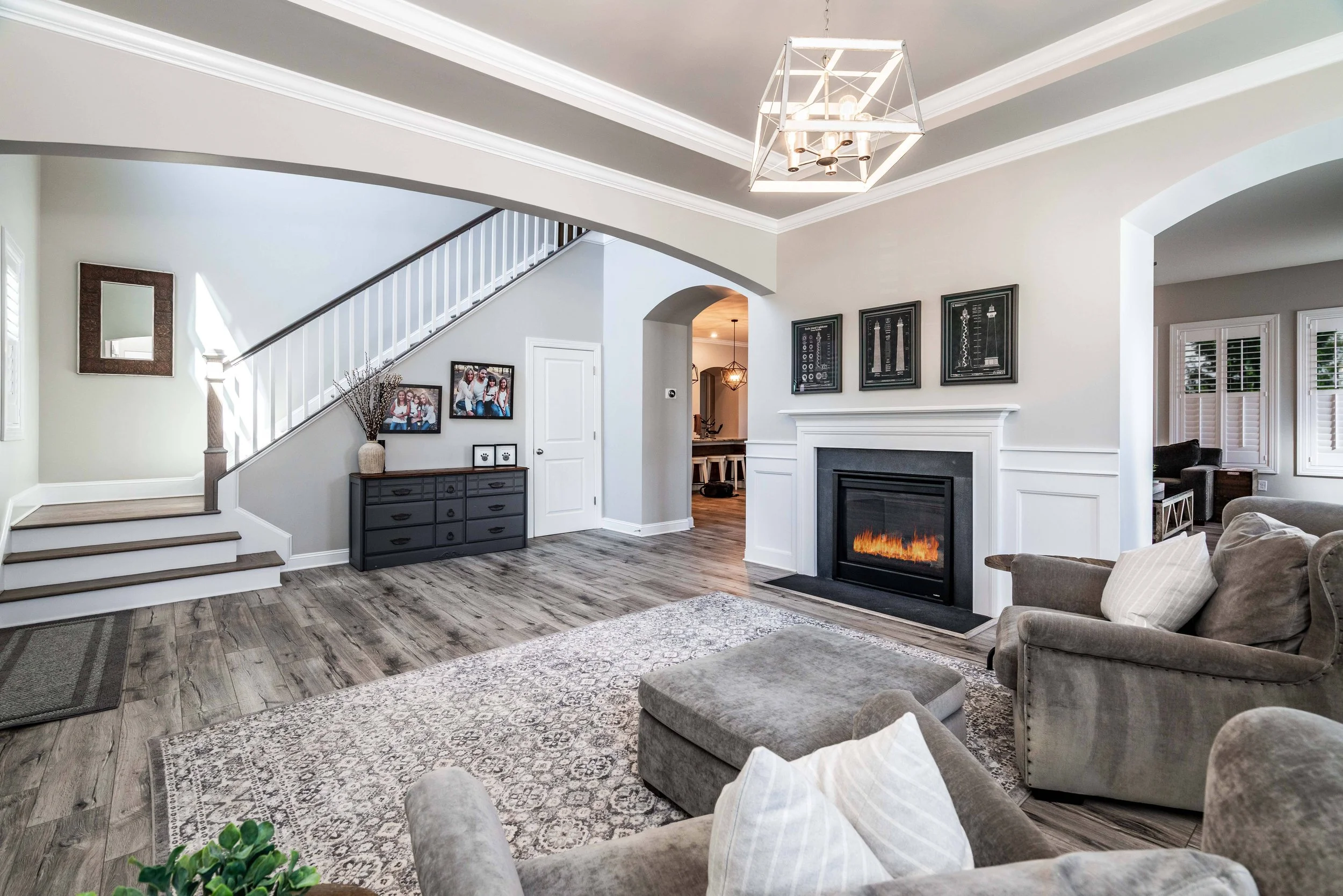 Spacious living room with a cozy fireplace, gray armchairs, and a patterned rug. Elegant staircase and chandelier, creating a warm, inviting ambiance.