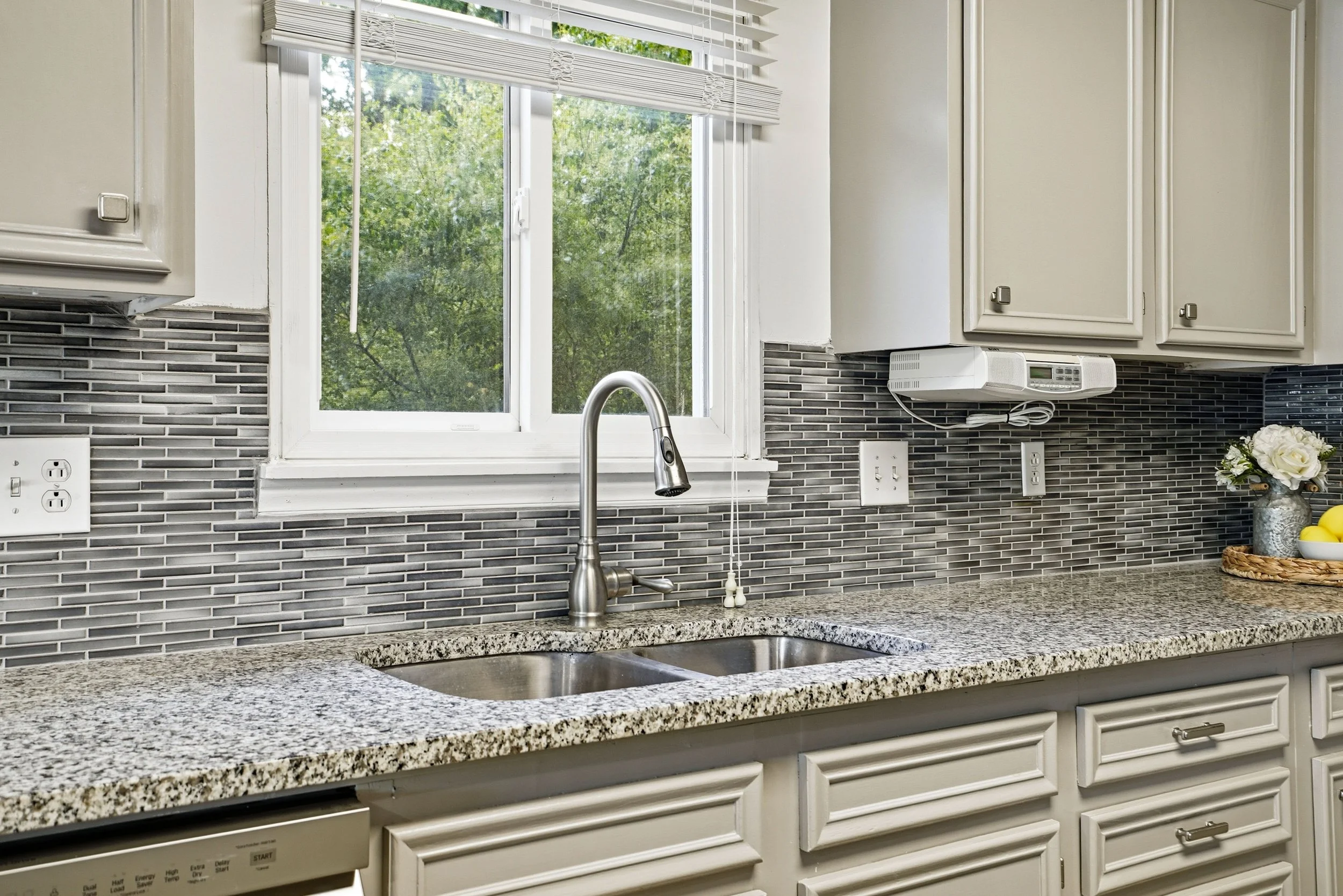 A modern kitchen featuring a gray and silver mosaic backsplash, dual stainless steel sinks, and a polished granite countertop. A large window above the sink provides natural light, revealing greenery outside.