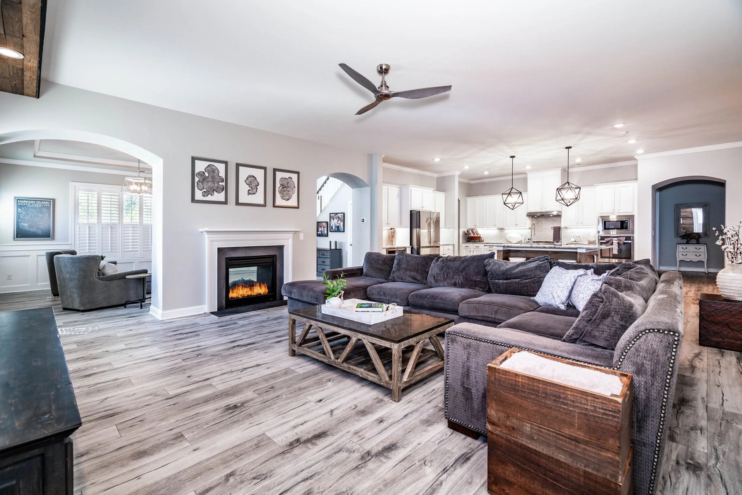 Spacious modern living room with a large gray sectional sofa, wooden coffee table, and electric fireplace. Open layout leads to a bright kitchen with white cabinets.