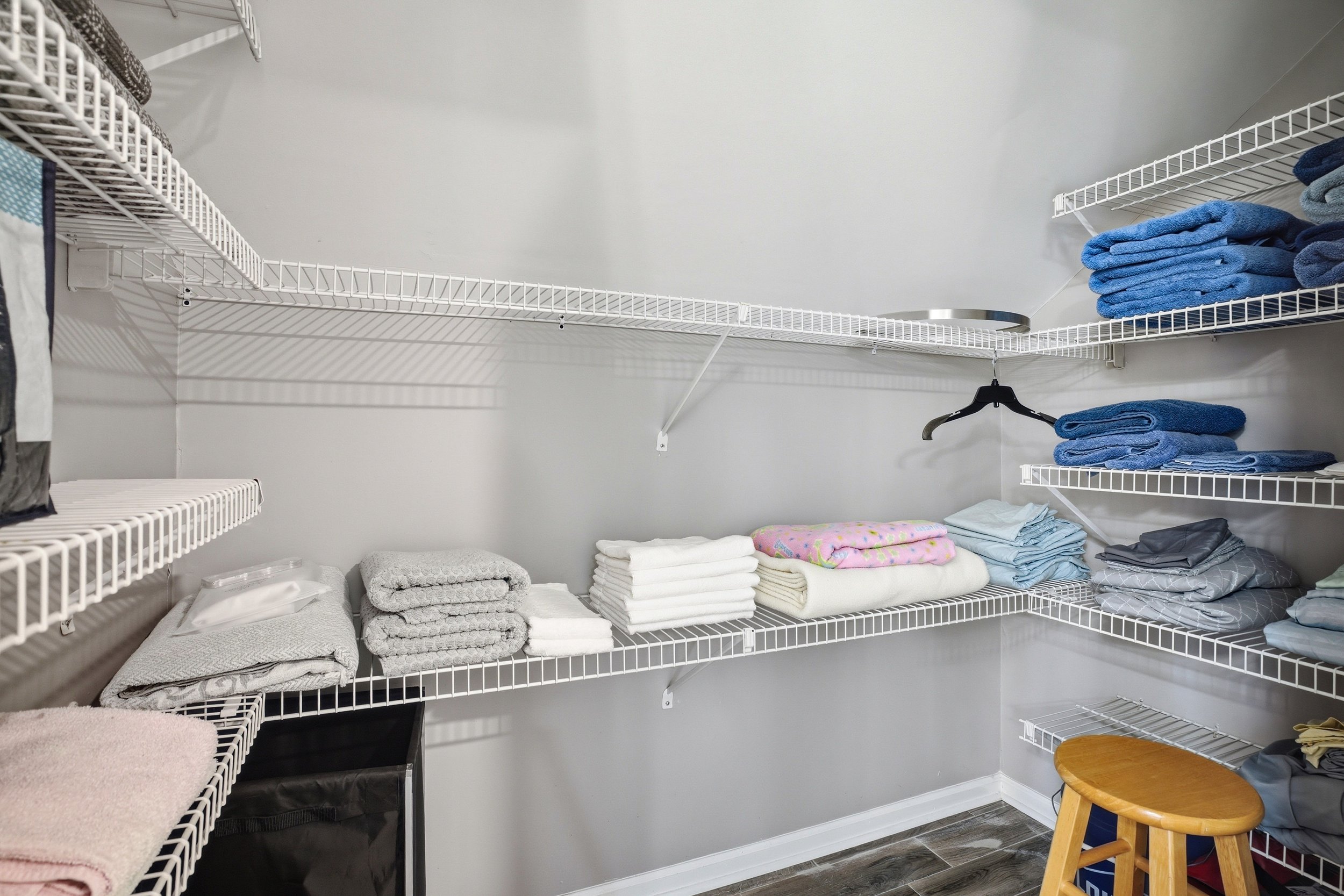 Walk-in closet with organized wire shelving holding neatly stacked towels, folded bedsheets, and blankets, with a wooden stool on the dark tile floor below.