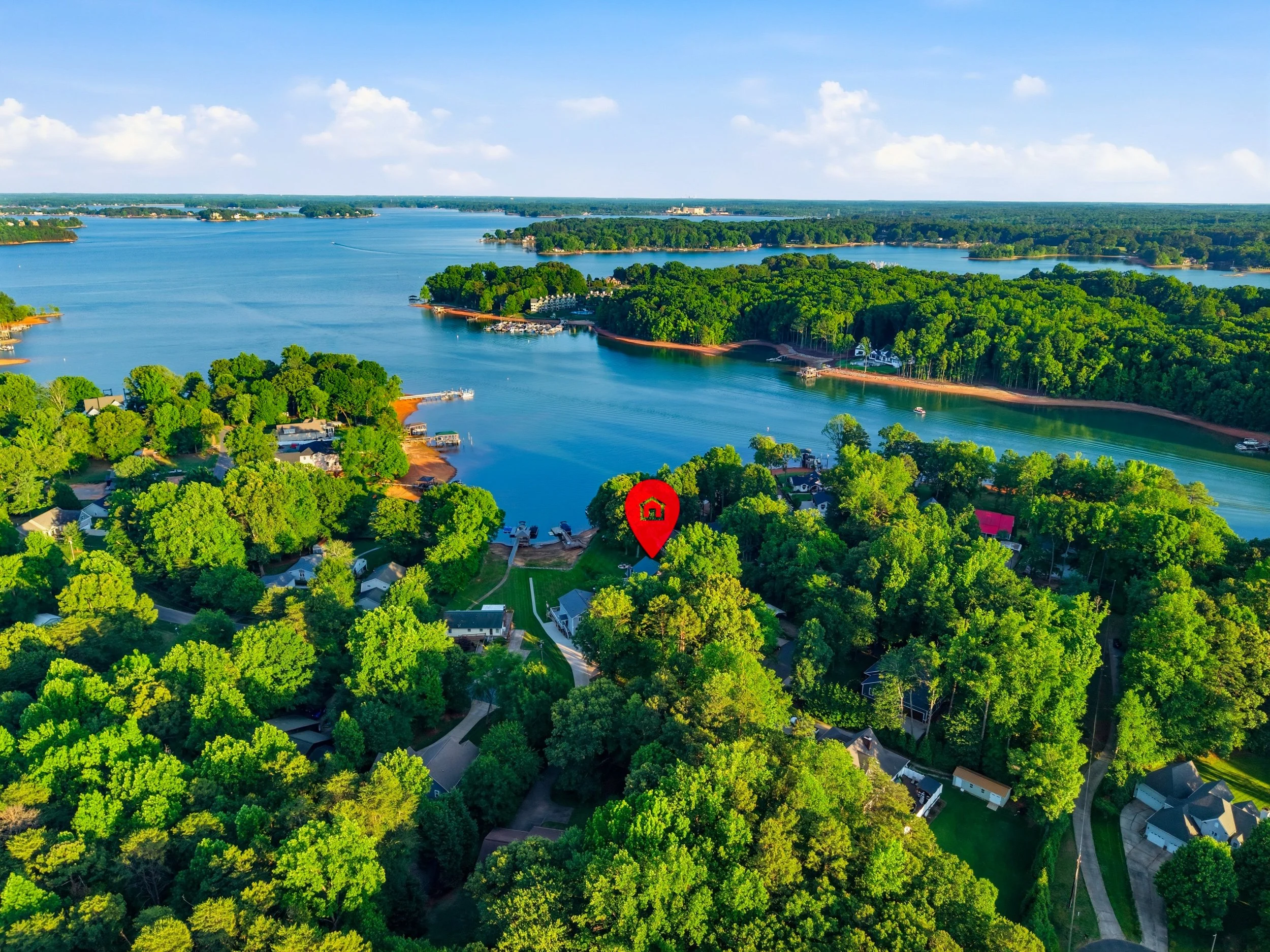 An aerial view of a lake surrounded by dense green forests and residential homes, with a red location pin marking a specific house near the water.