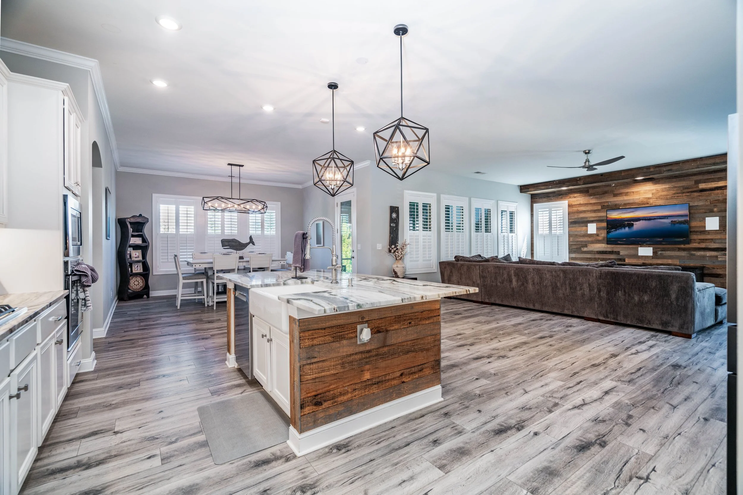 Open-concept living space with a modern kitchen, dining area, and living room. Features white cabinets, wood accents, geometric pendant lights, and a cozy brown sofa facing a TV on a wooden accent wall. Bright and inviting atmosphere.