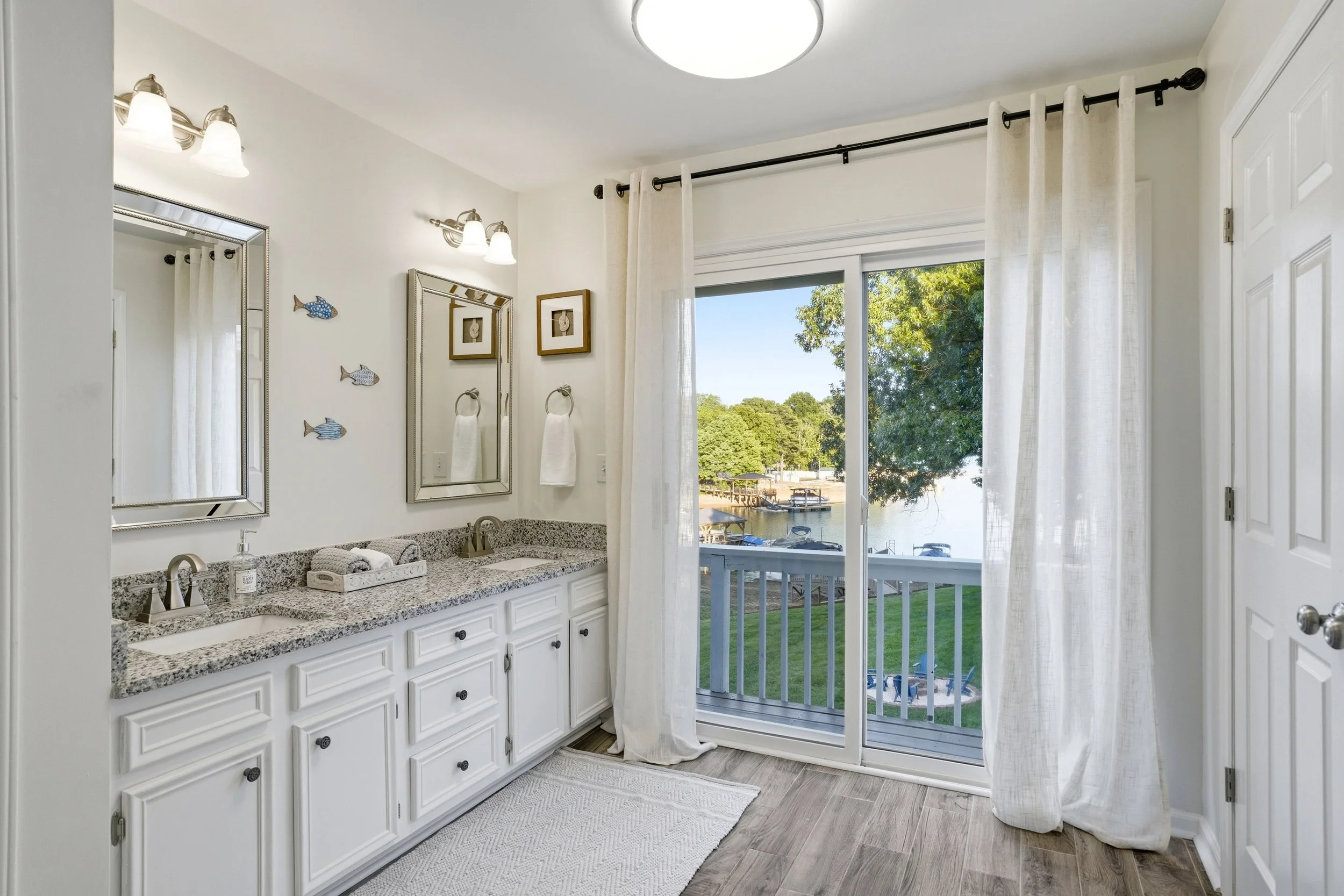 Bright bathroom featuring a double vanity with granite countertops, dual mirrors, and a sliding glass door opening to a deck overlooking a lake.