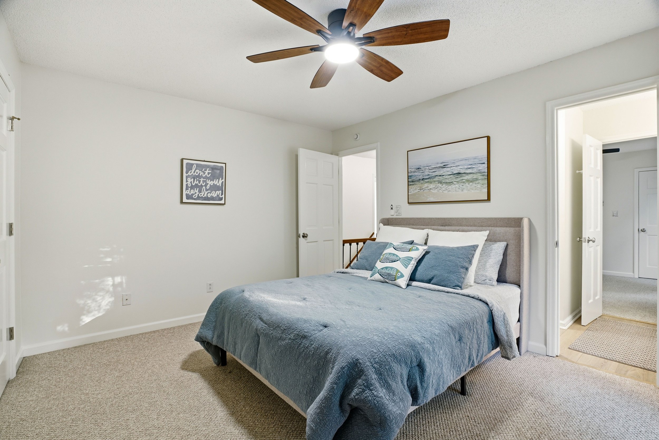 A carpeted bedroom with white walls, featuring a bed with a gray comforter, a ceiling fan, and doors leading to a hallway and bathroom.