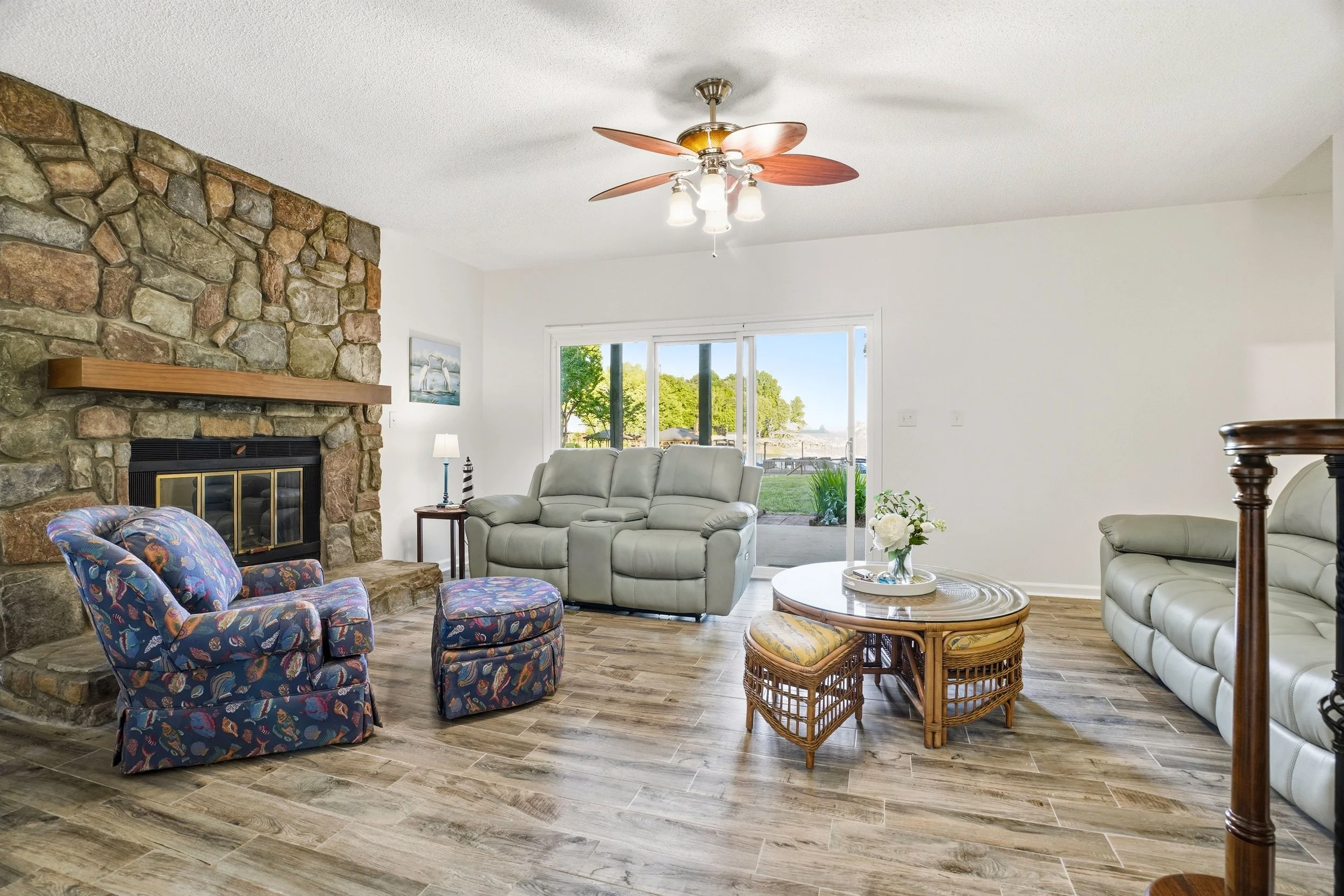 Spacious living room featuring wood-plank flooring, a prominent stone fireplace, two grey leather couches, a floral-patterned armchair with an ottoman, and a glass-topped coffee table, with a sliding glass door opening to a view of trees outside.