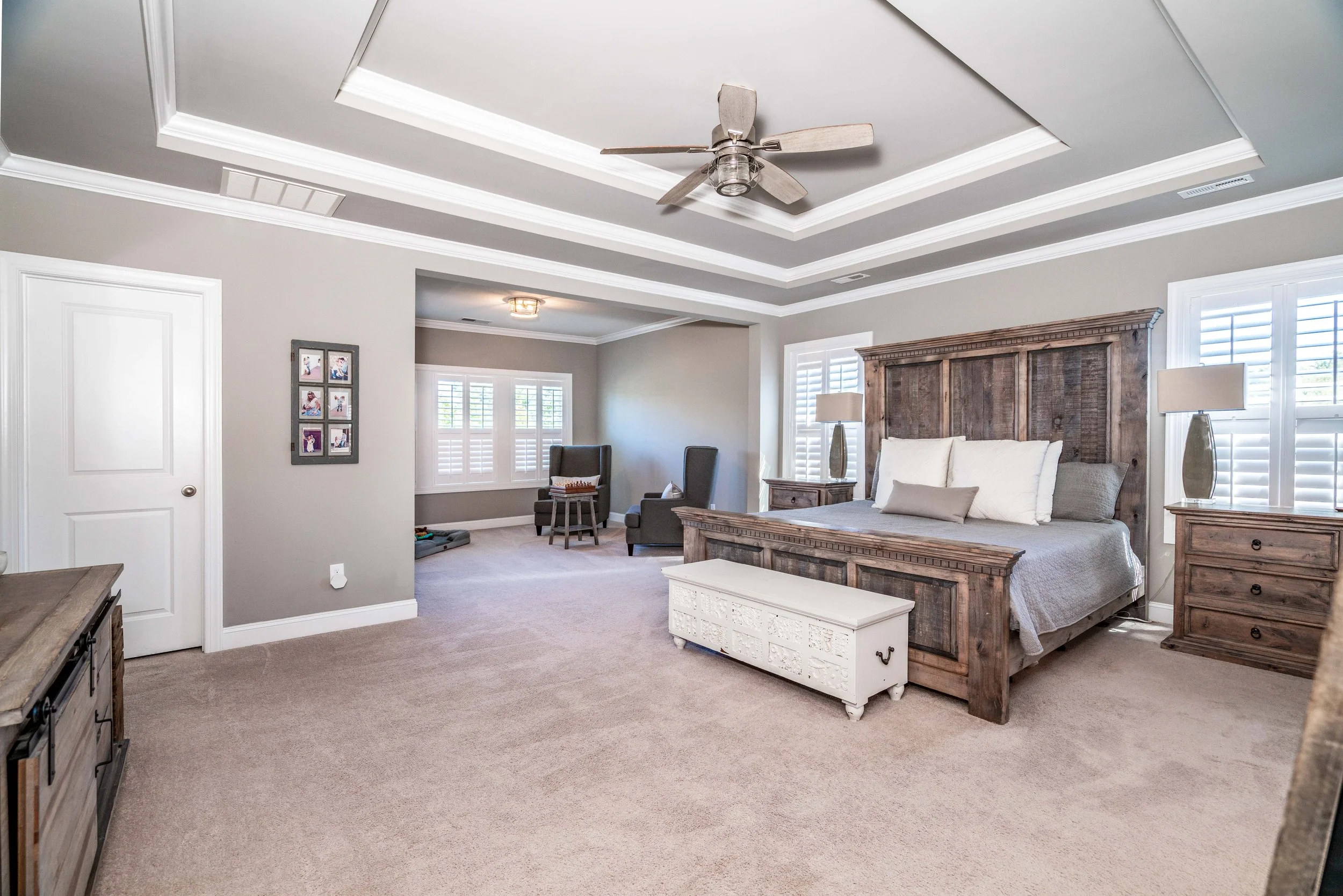 Spacious bedroom with rustic wood furniture, gray walls, and tray ceiling. Includes a large bed, chest, armchair, and shuttered windows, conveying a cozy, modern style.