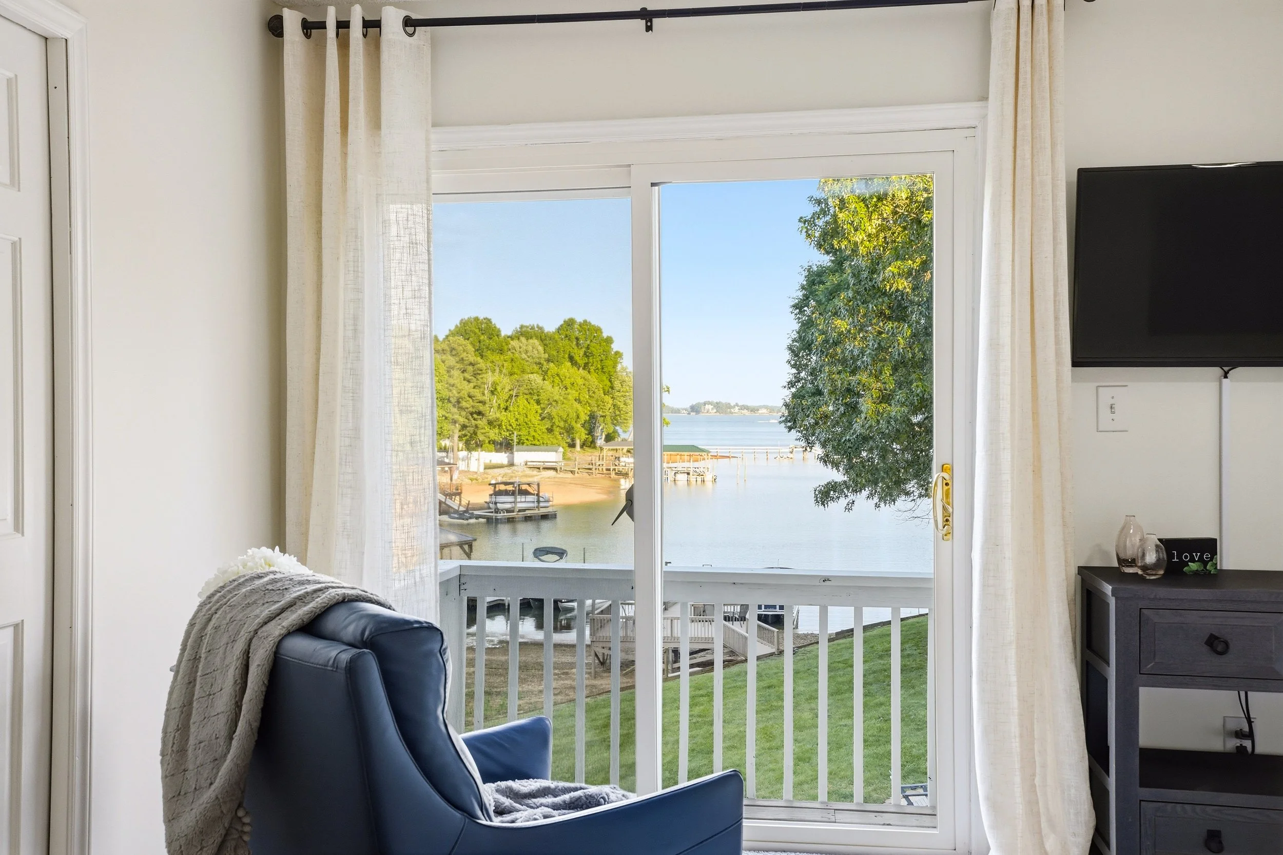 A sliding glass door opens to a balcony overlooking a lake with docks and trees, viewed from a room with a blue armchair and TV.
