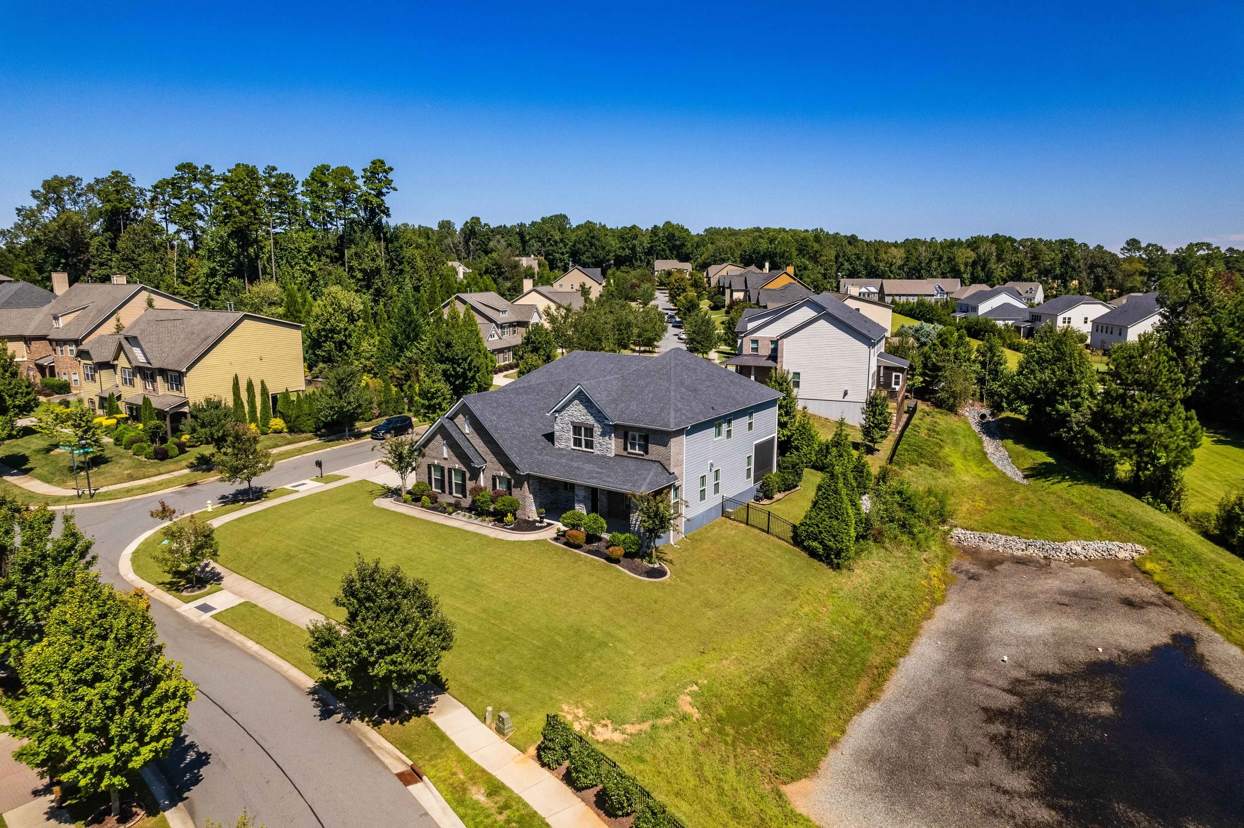 Aerial view of a suburban neighborhood with green lawns and trees. A large, modern house is centered, surrounded by neatly trimmed grass and a curved road, under a clear blue sky.