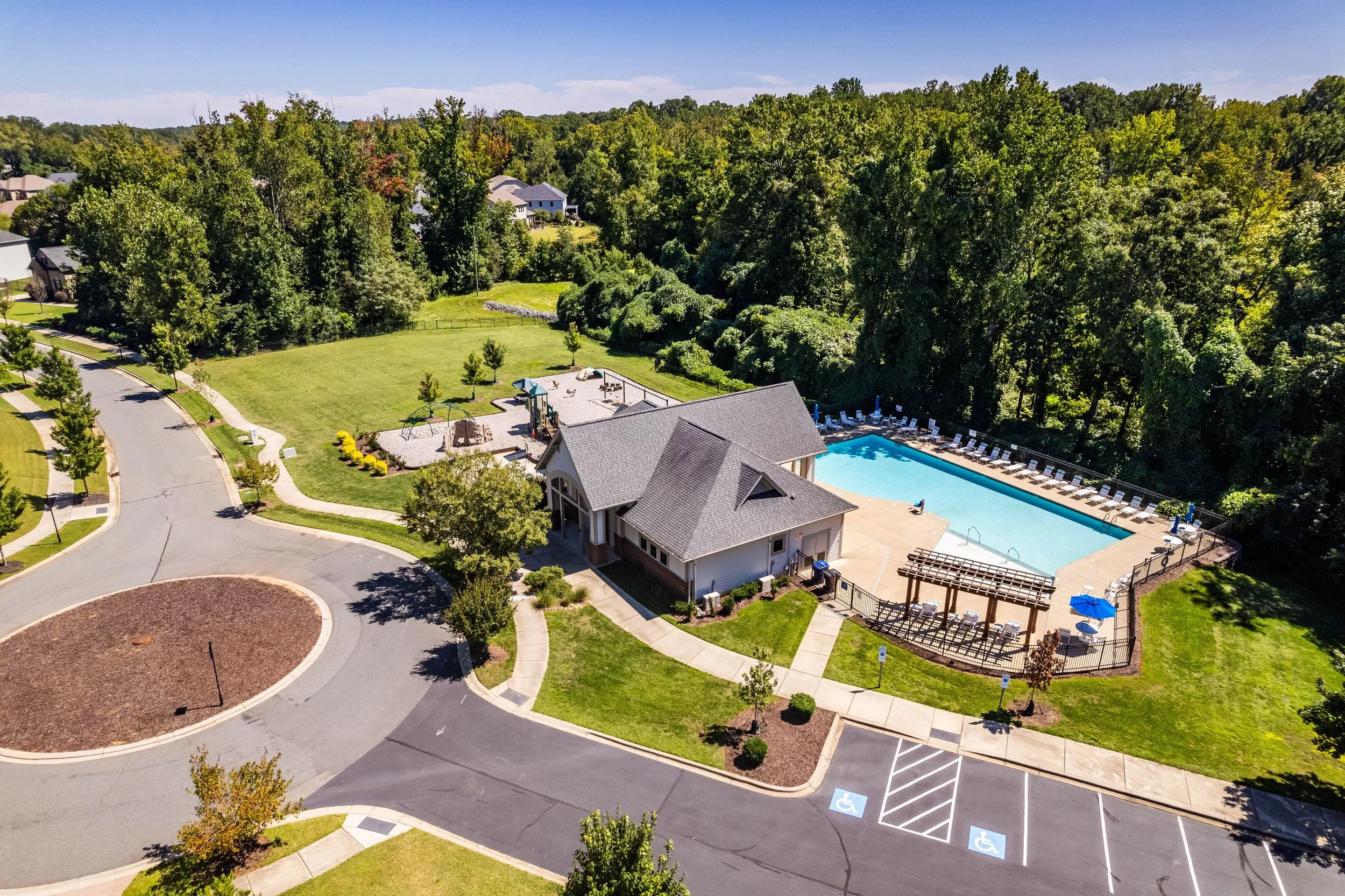 Aerial view of a community park with a rectangular swimming pool next to a clubhouse. Lush green trees surround the area, creating a serene atmosphere.