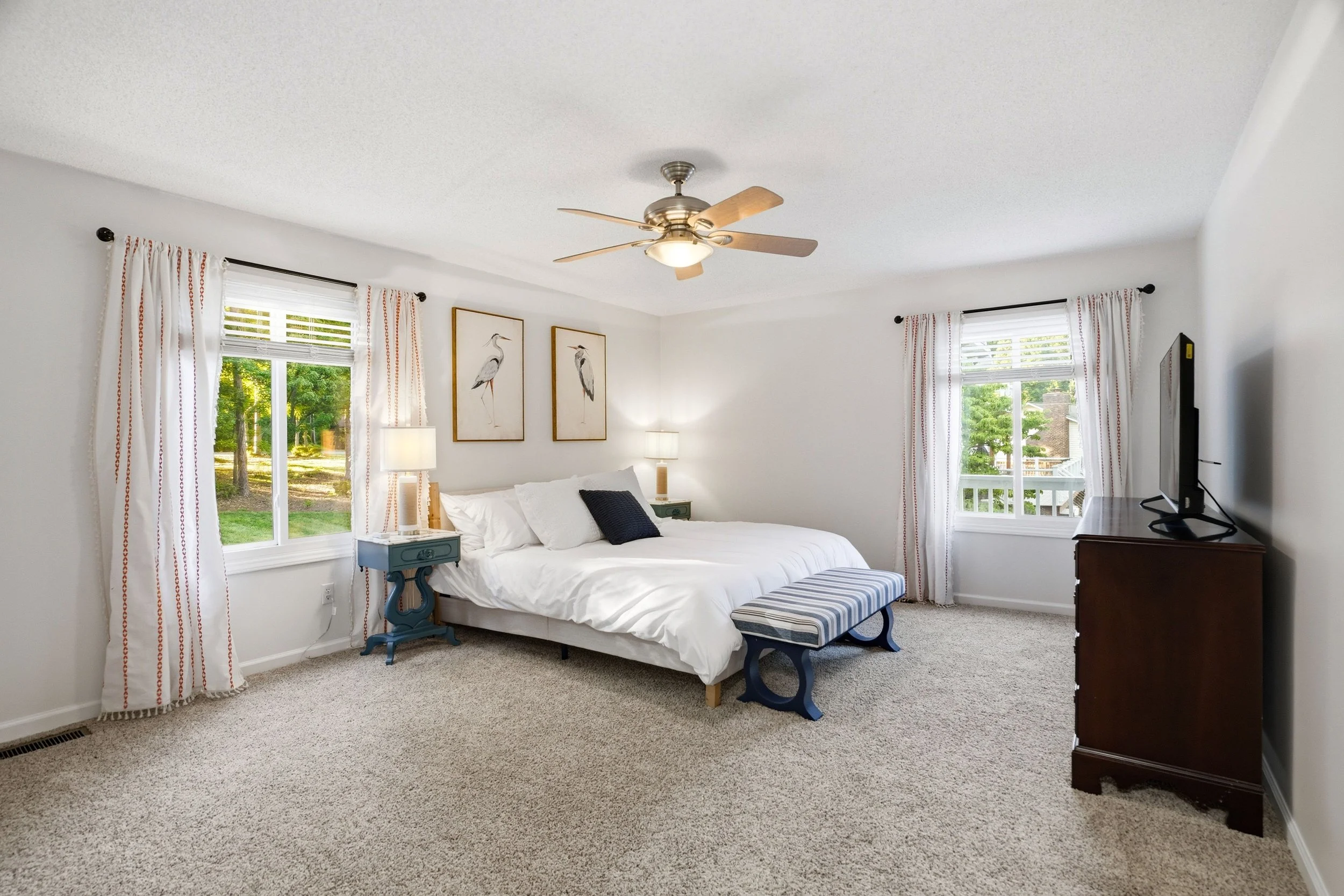 A spacious bedroom features a king-size bed with white bedding and a striped bench at the foot. Two windows, adorned with light curtains, allow natural light to brighten the room. A dresser and nightstand are positioned next to the bed.