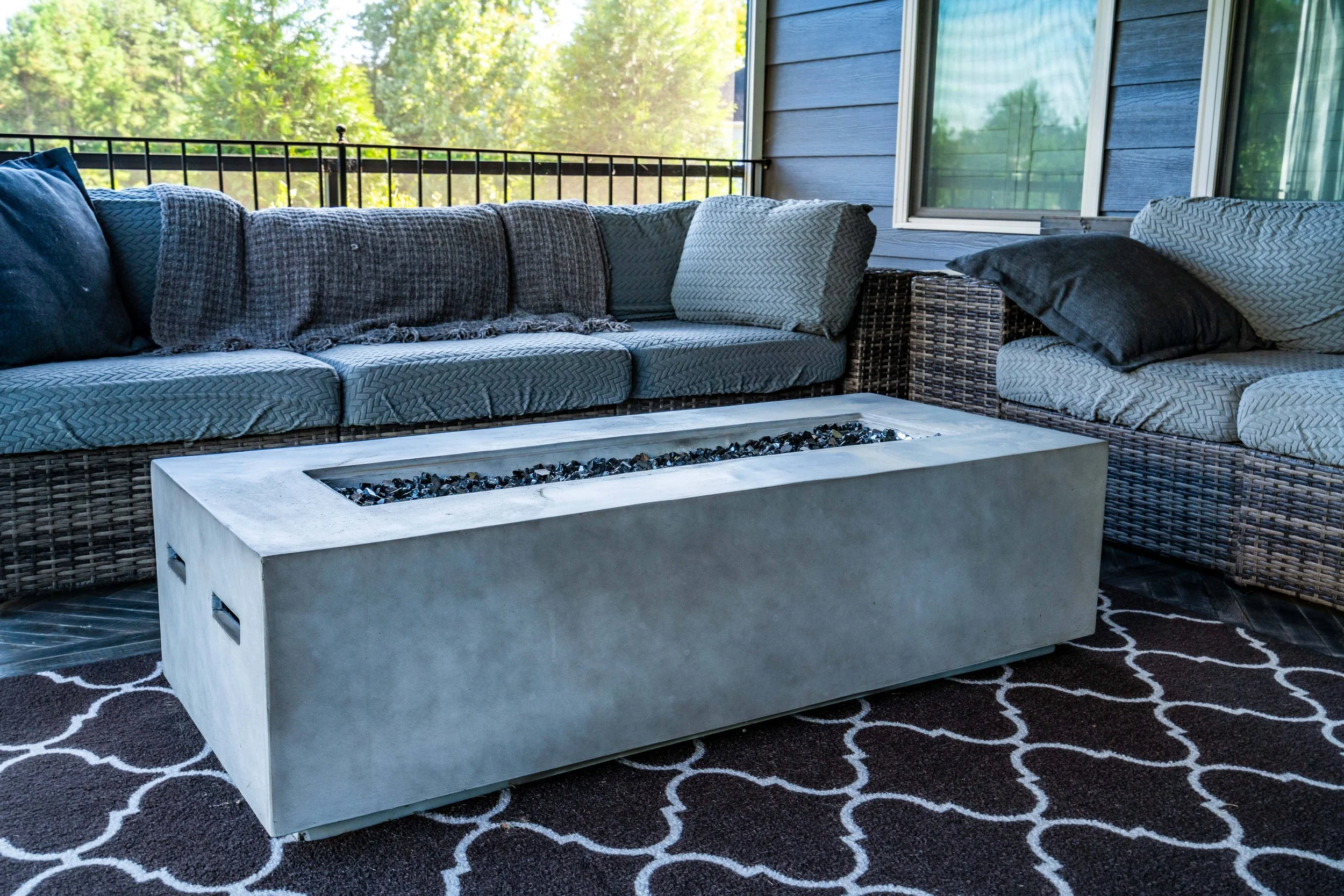 Stylish patio with gray wicker sofas, colorful cushions, and a modern rectangular fire pit on a patterned rug. Wood and foliage visible through windows.