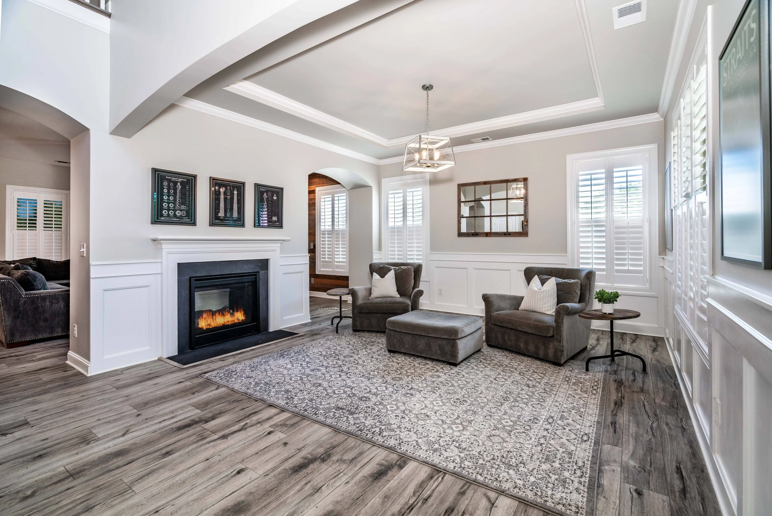 Modern living room with a cozy fireplace, two gray armchairs, an ottoman, and a patterned rug. Sunlight filters through white shutters, creating a serene ambiance.