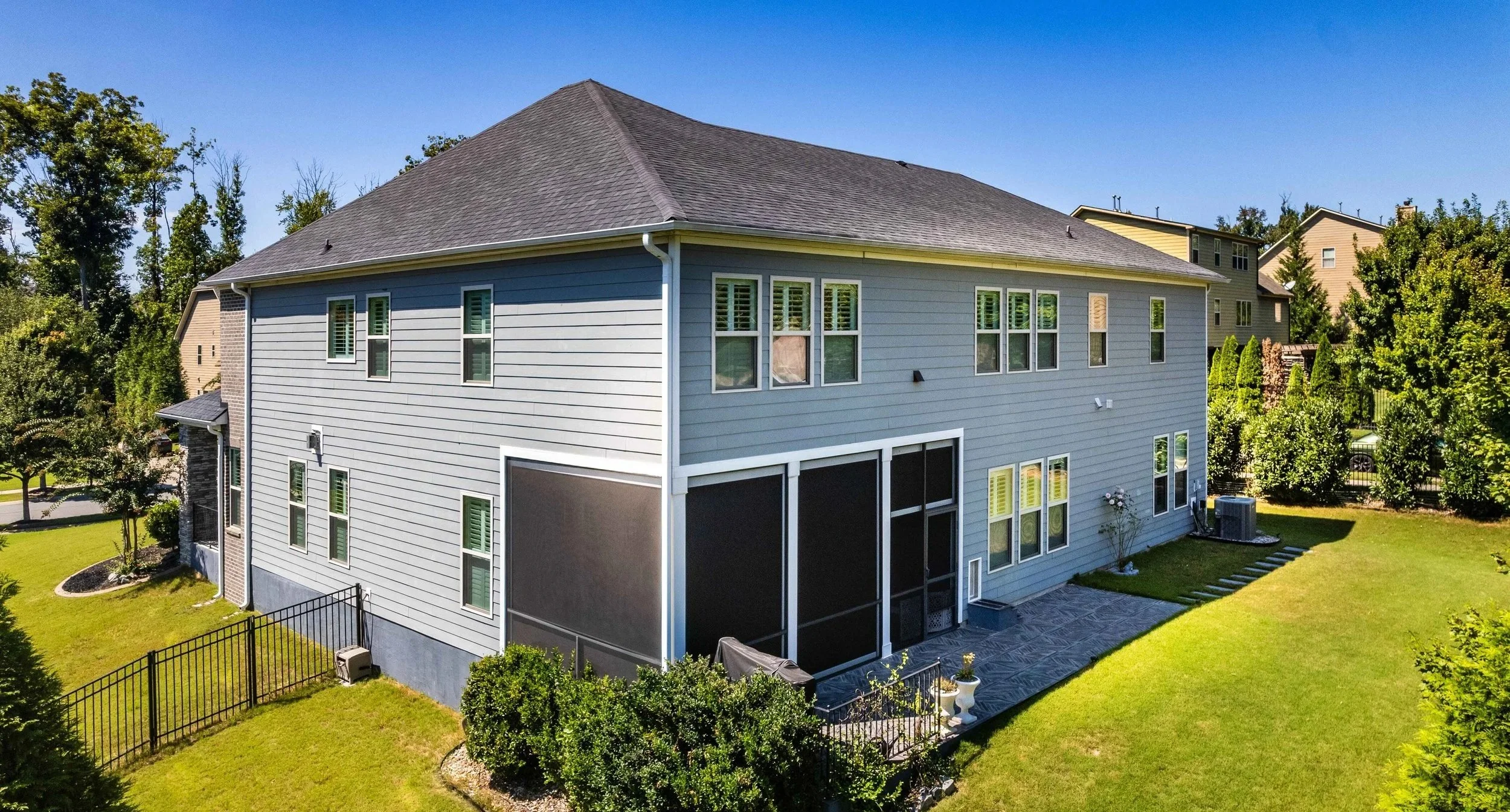 Large modern gray house with a dark roof, screened patio, and expansive green lawn. Surrounded by trees and shrubs under a clear blue sky. Peaceful.