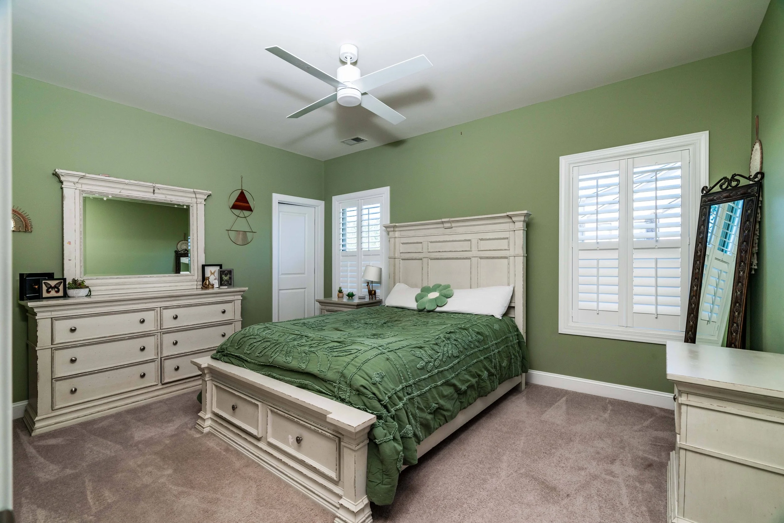 A cozy bedroom features green walls, a white bed with a green comforter, and matching white furniture. A ceiling fan hangs above, creating a serene atmosphere.