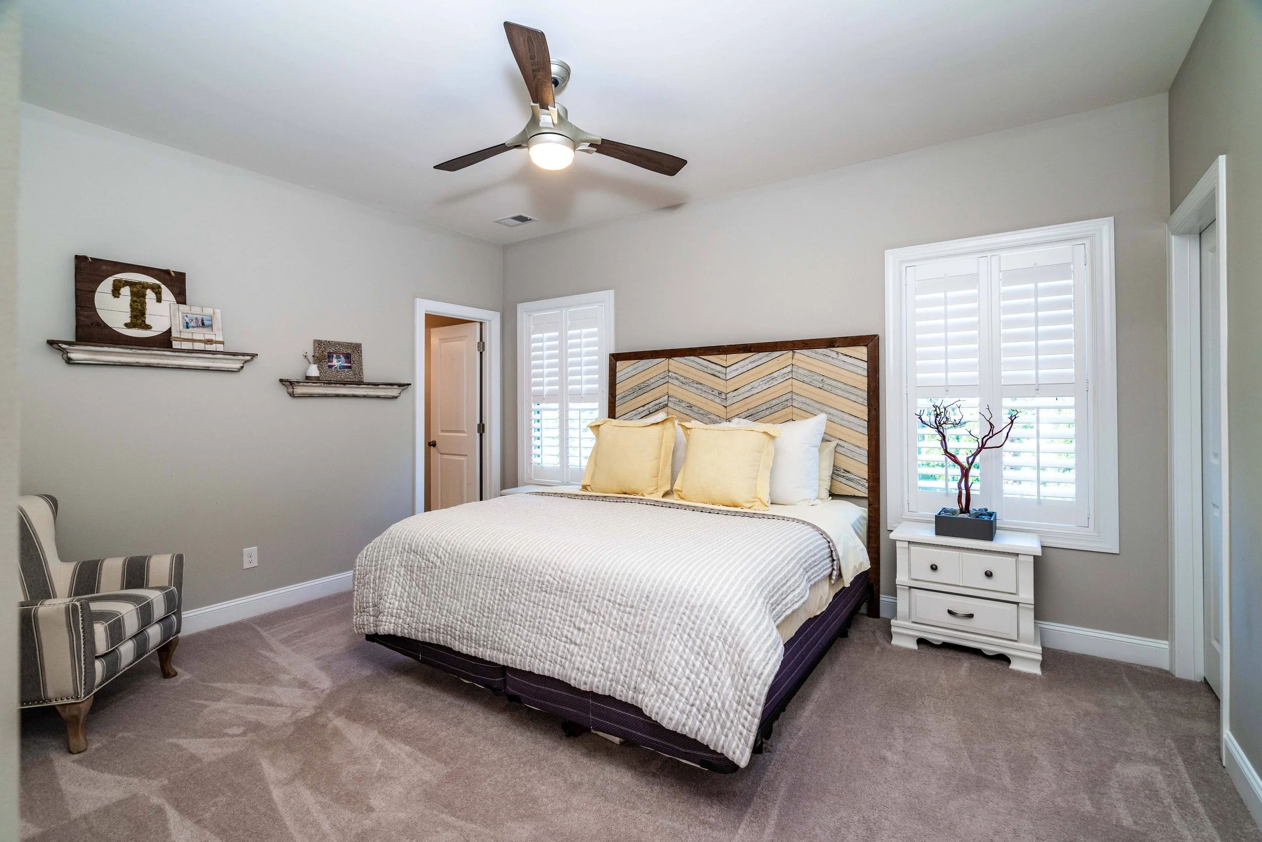 A cozy bedroom with a large bed featuring white bedding and two yellow pillows. Beige walls, striped armchair, and wooden accents create a calming vibe.