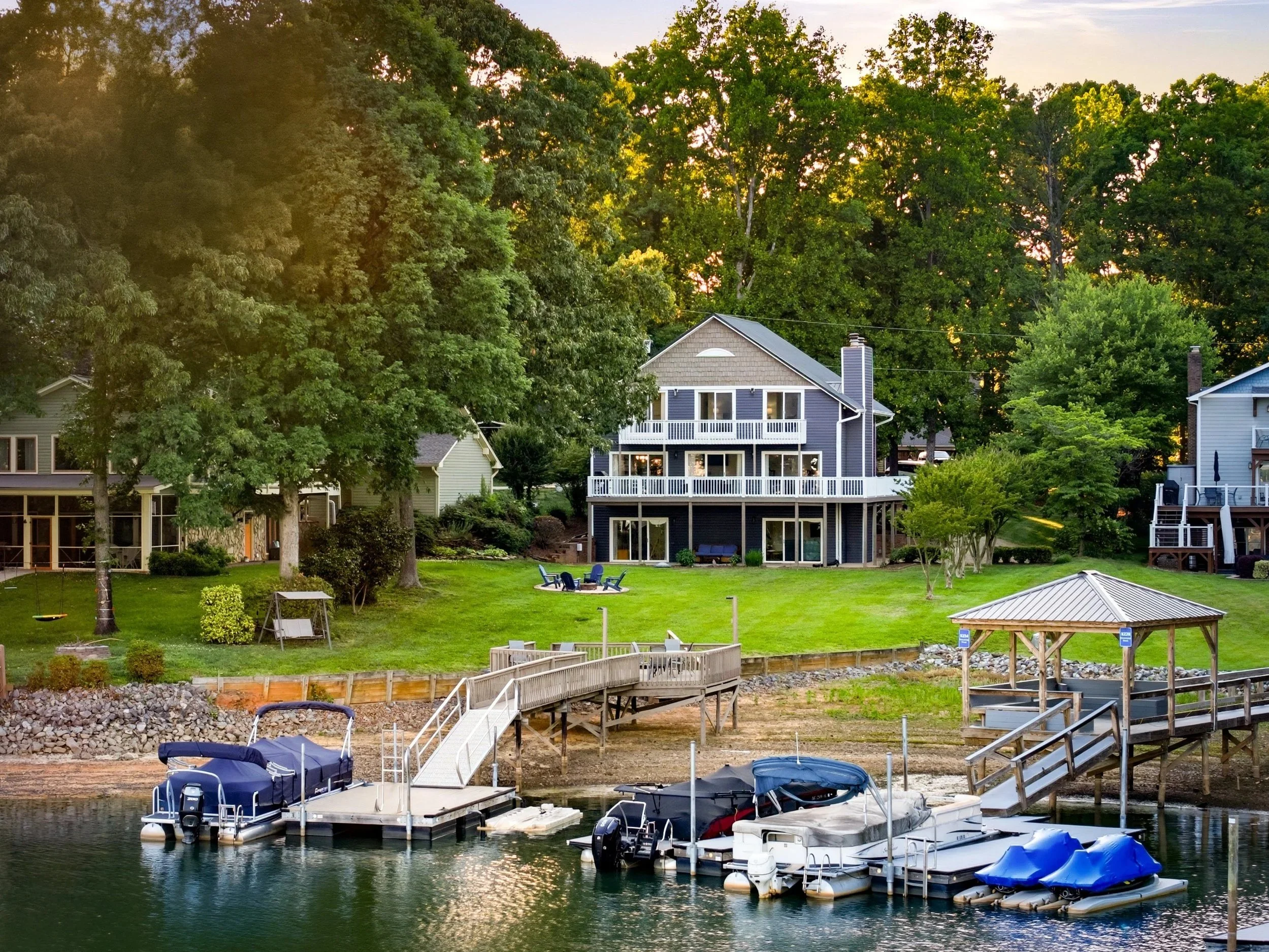 A multi-story blue house with white balconies sits on a grassy hill by a lake, featuring boat docks with pontoon boats and jet skis in the water.