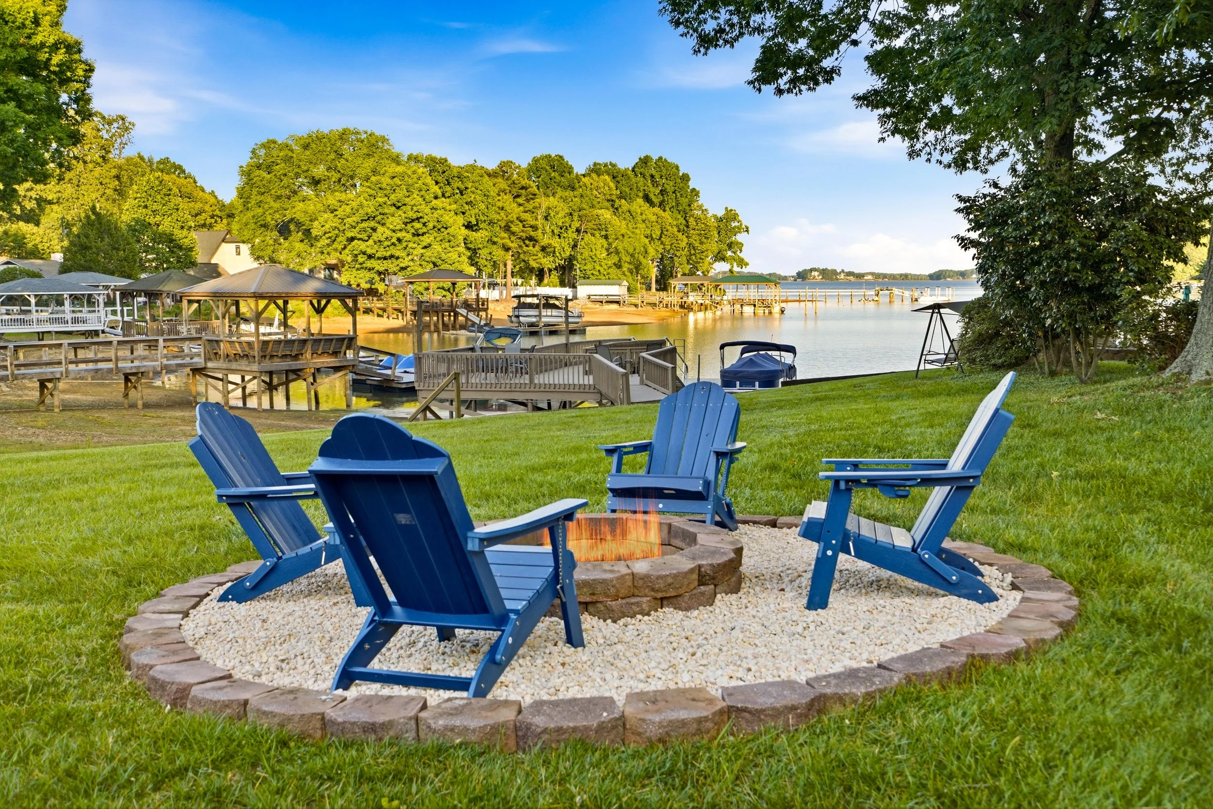 Four blue Adirondack chairs arranged around a circular stone fire pit filled with gravel on a grassy lawn overlooking a lake with docks and trees.