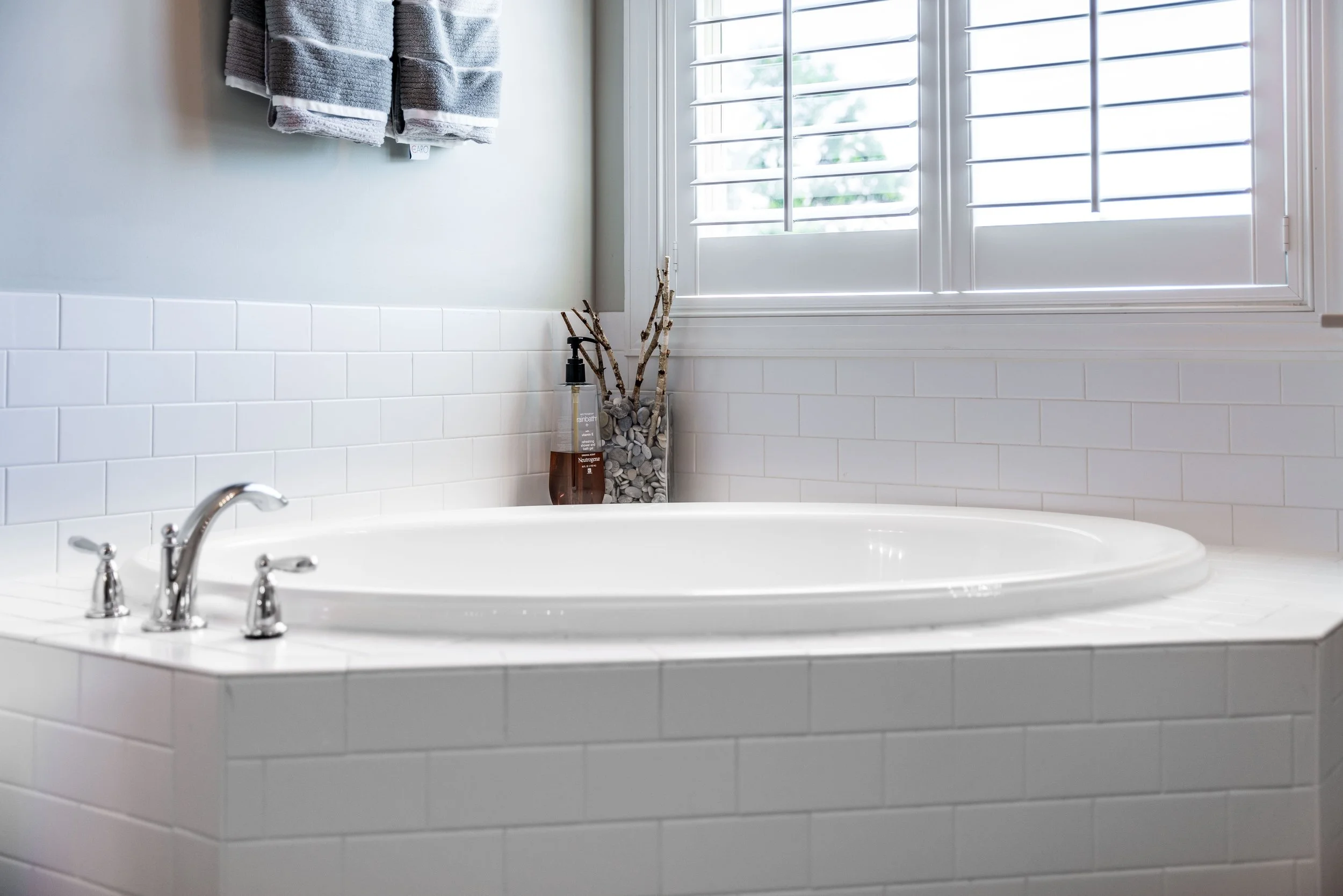 Elegant bathroom with a white corner bathtub, silver faucets, and white tiled walls. Sunlight filters through closed blinds. Relaxing and serene.