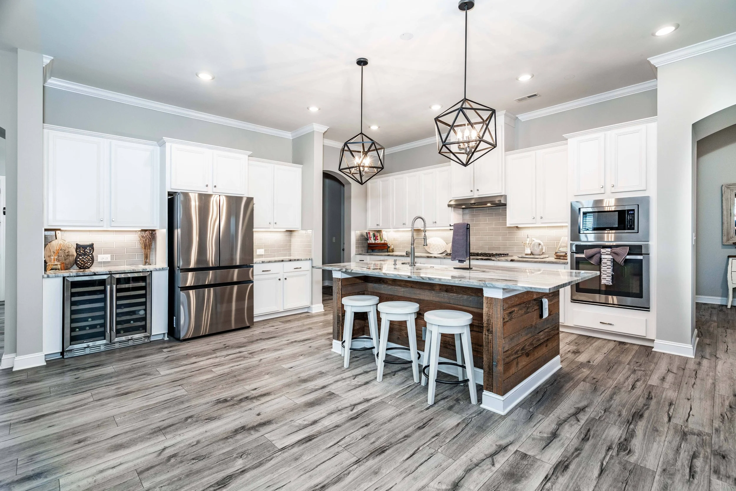 Modern kitchen with white cabinets, stainless steel appliances, and a wooden island with three stools. Geometric pendant lights add elegance. Bright and inviting.