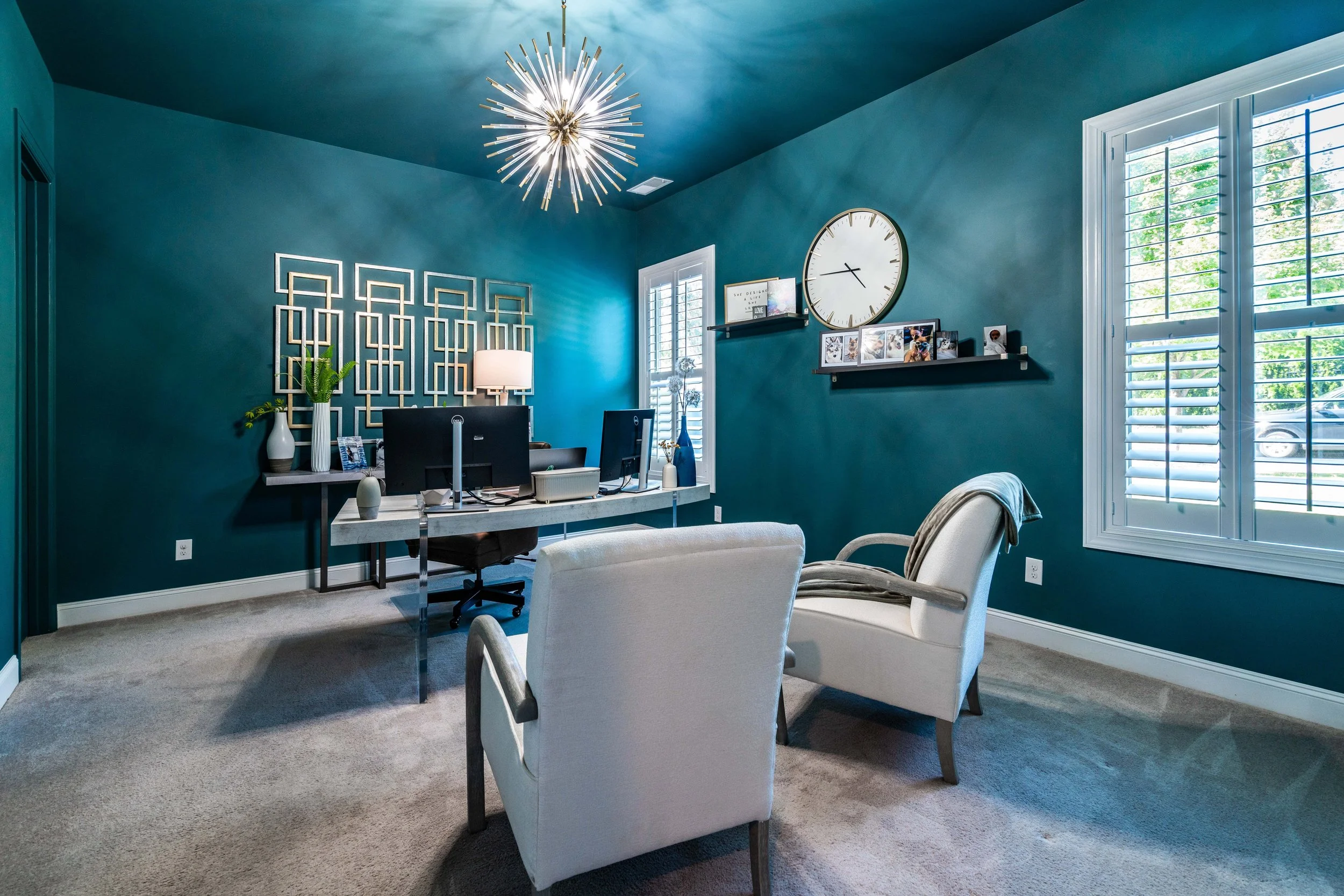 Stylish home office with teal walls, a modern starburst light fixture, large clock, desk with computer, white chairs, and window shutters. Cozy and elegant.