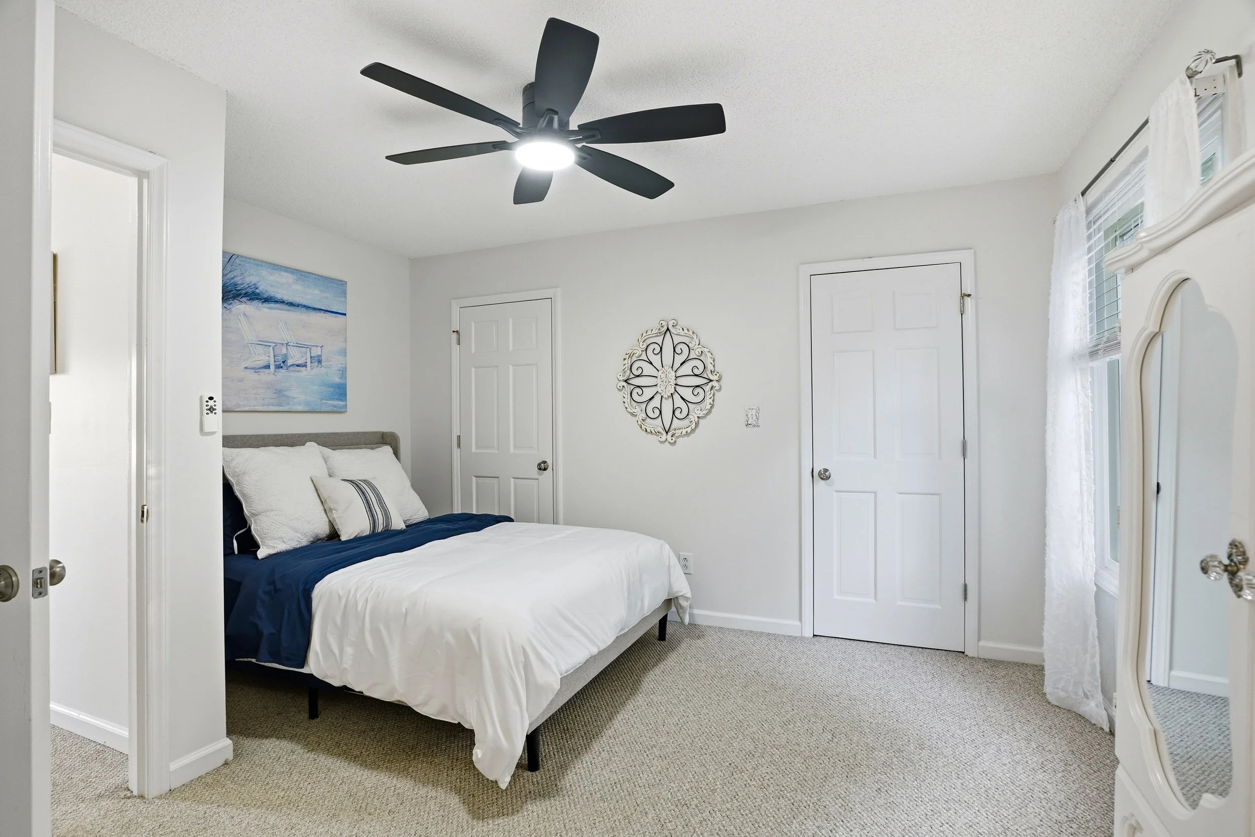 Bright, minimalist bedroom with white walls, a bed made with white and blue linens, a dark ceiling fan, and decorative wall art.