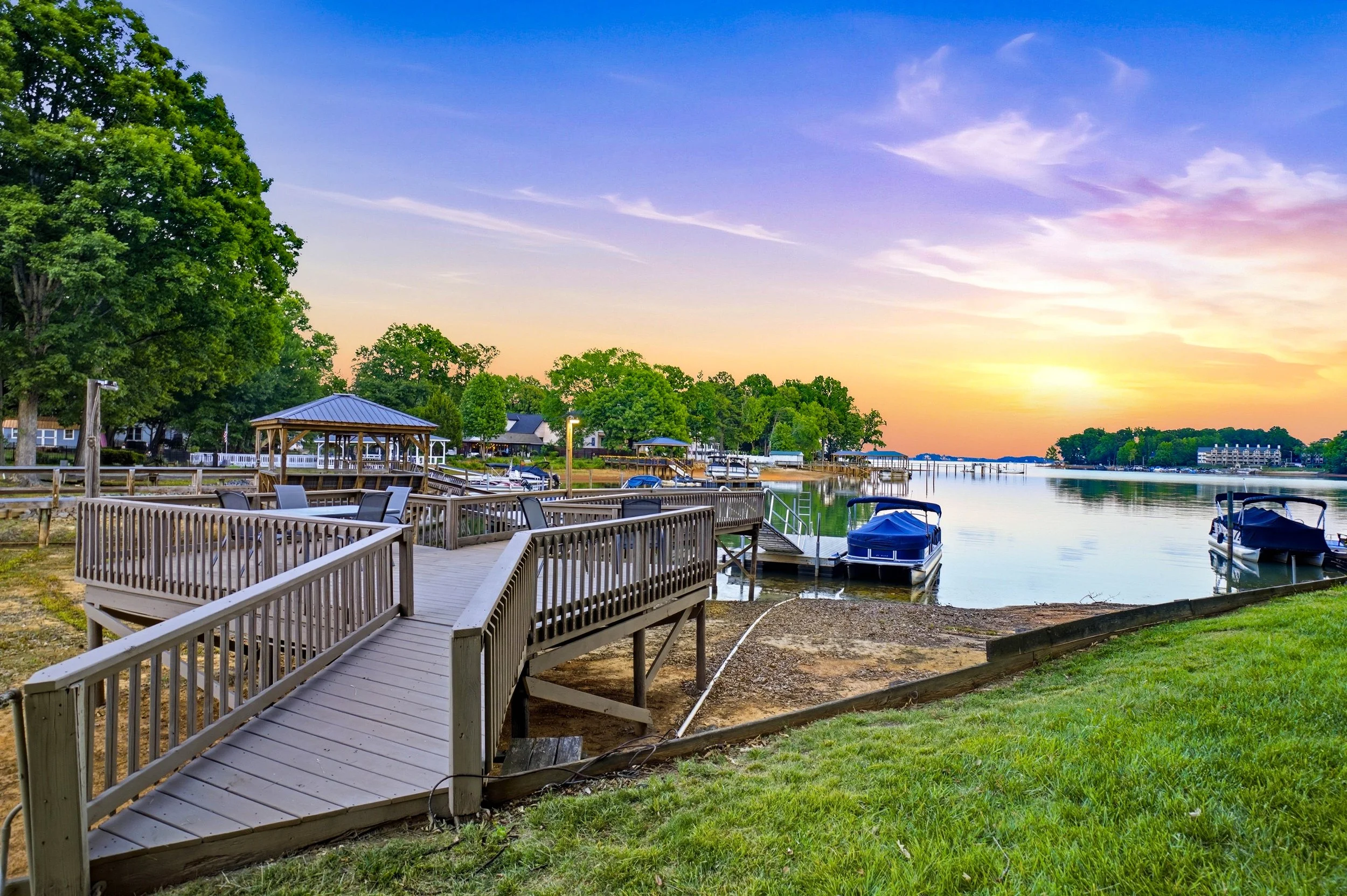 A wooden deck with railings overlooks a calm lake at sunset, featuring a gazebo, docked pontoon boats, and a grassy shoreline under a colorful sky.