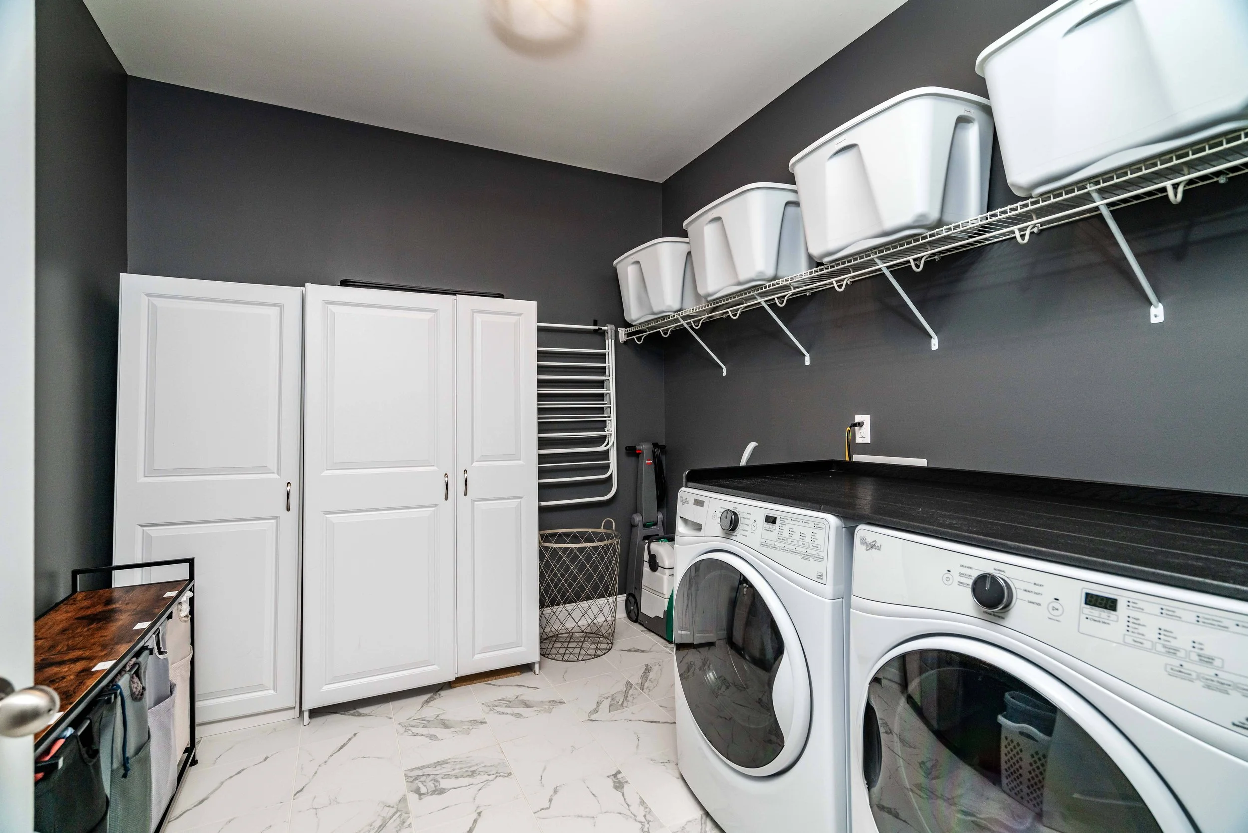 A clean, modern laundry room with dark gray walls and marble flooring. It features a washer and dryer, white storage bins on shelves, and a drying rack.