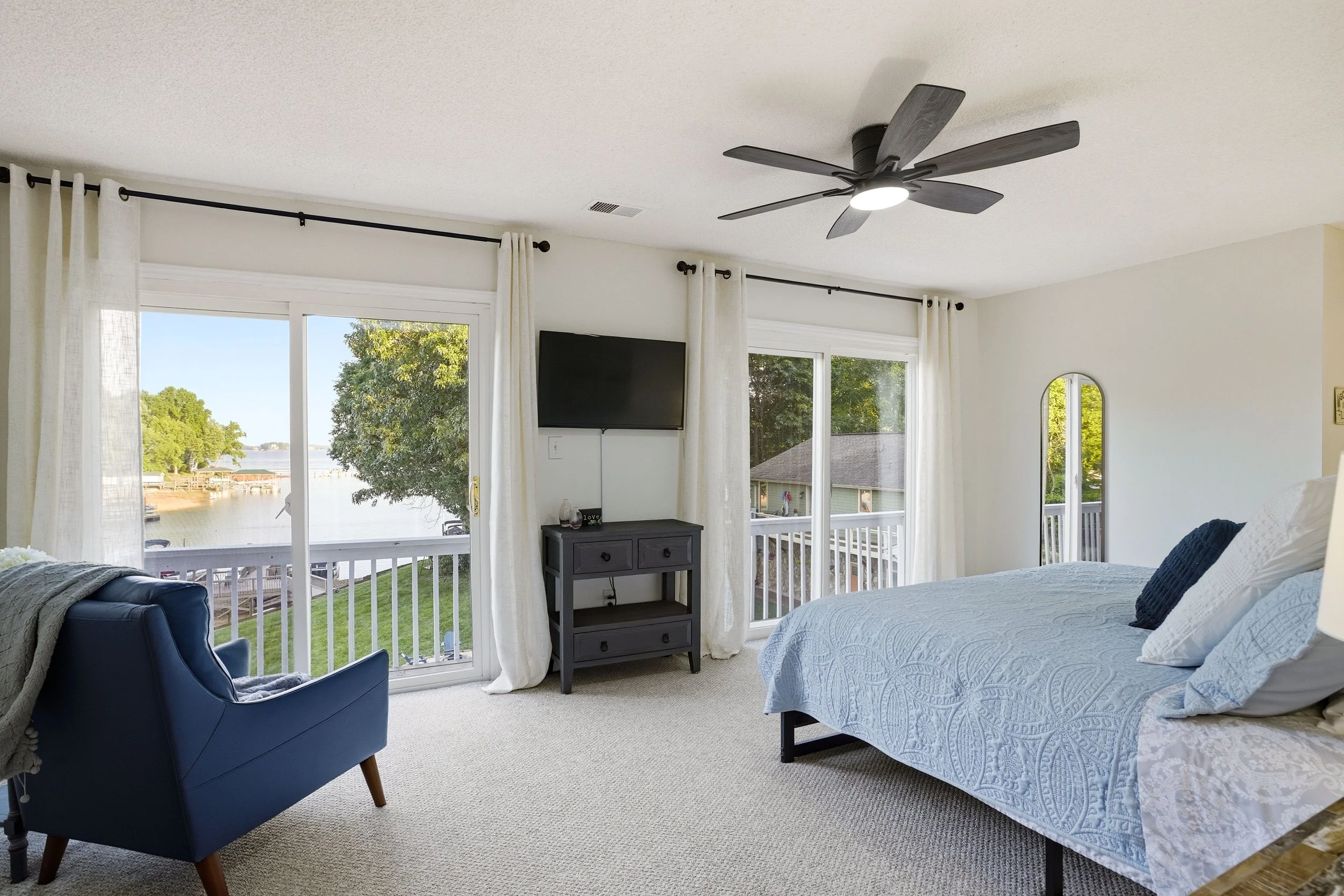 Bright bedroom featuring a large bed, blue armchair, and sliding glass doors that open to a scenic lake view with trees along the shore.