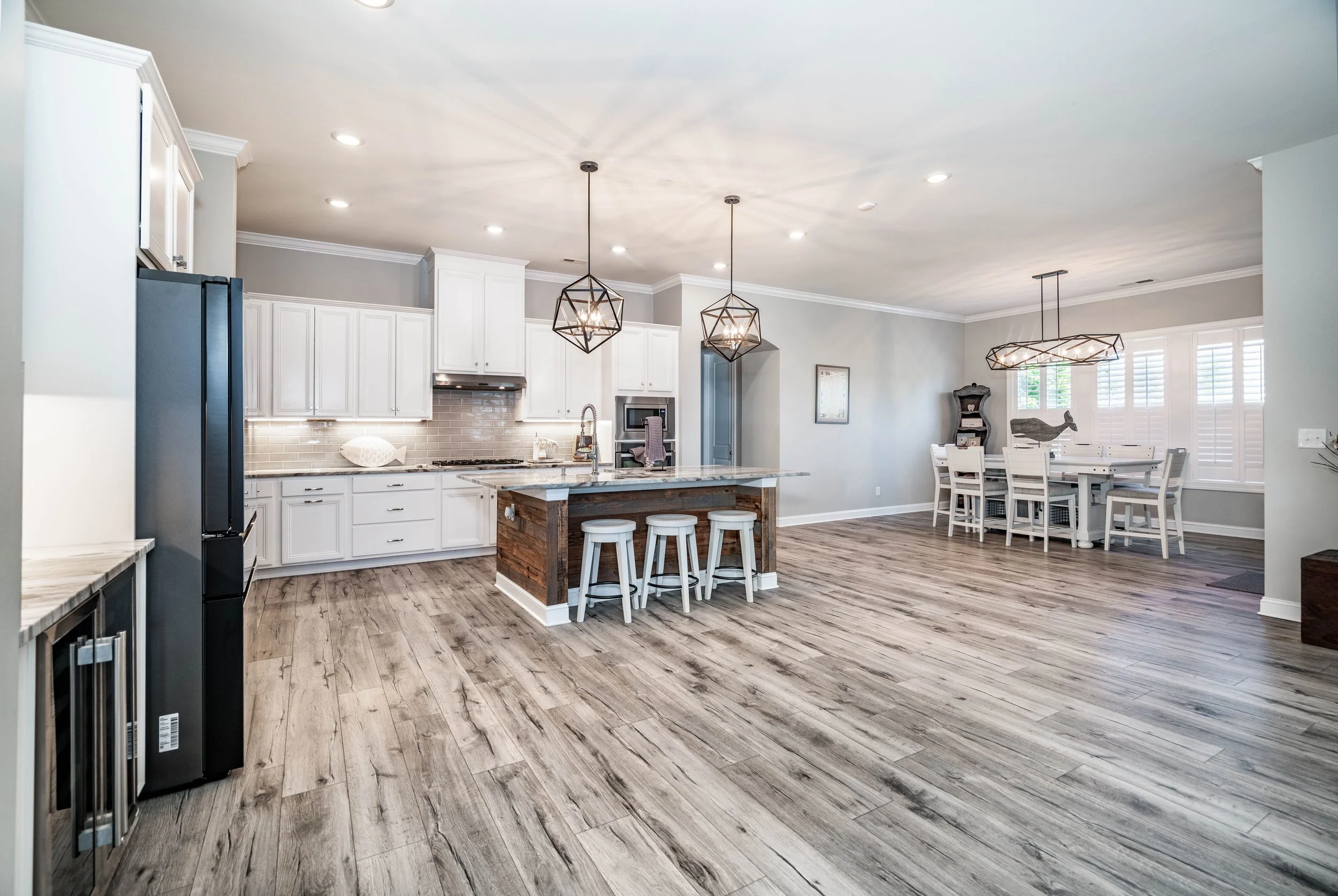 Spacious, modern kitchen with white cabinets, wooden island, and hanging geometric lights. Dining area with white table and natural light from windows.