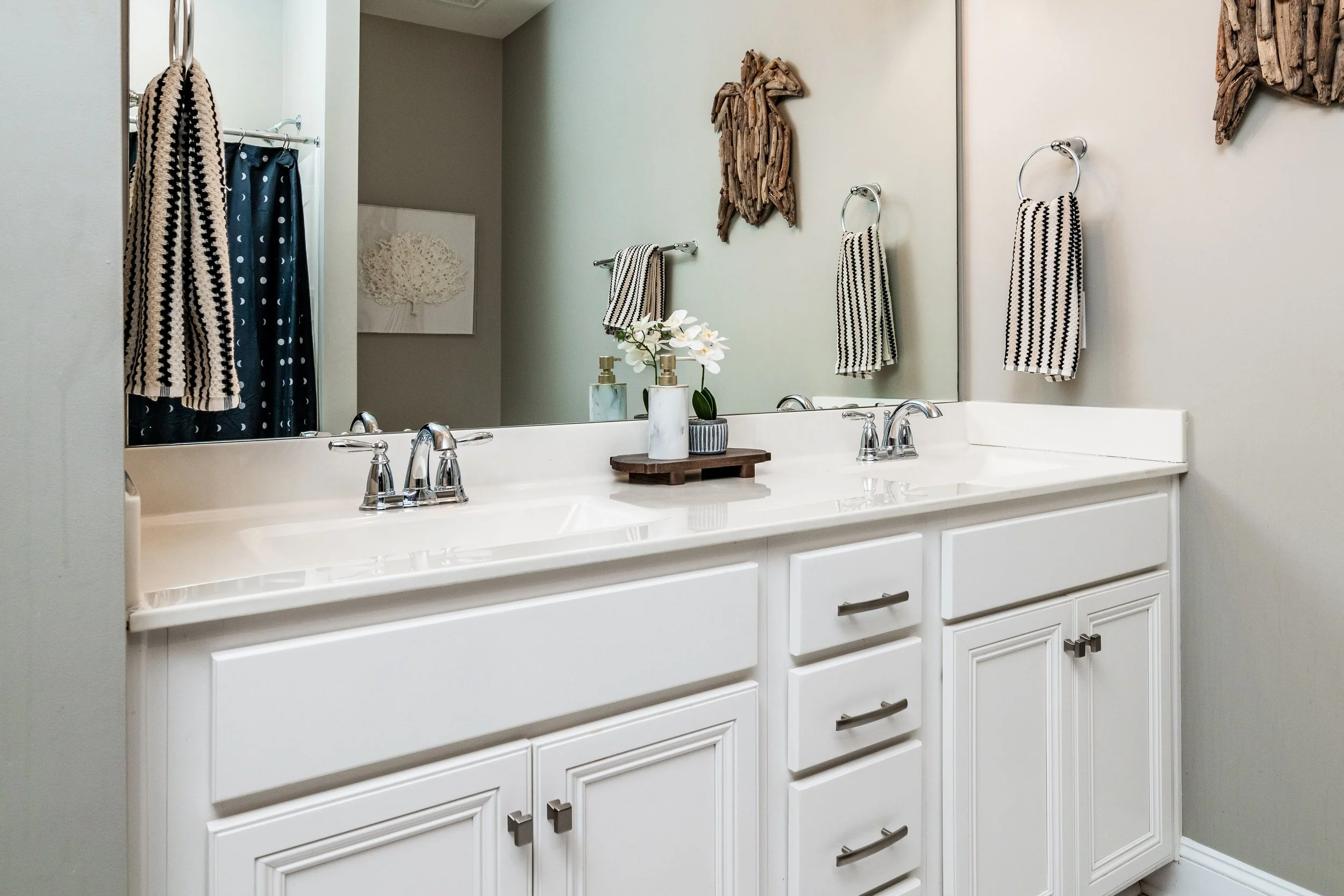 Elegant bathroom with a double vanity featuring white cabinets and silver fixtures. A wooden decor piece and hand towels enhance a serene ambiance.