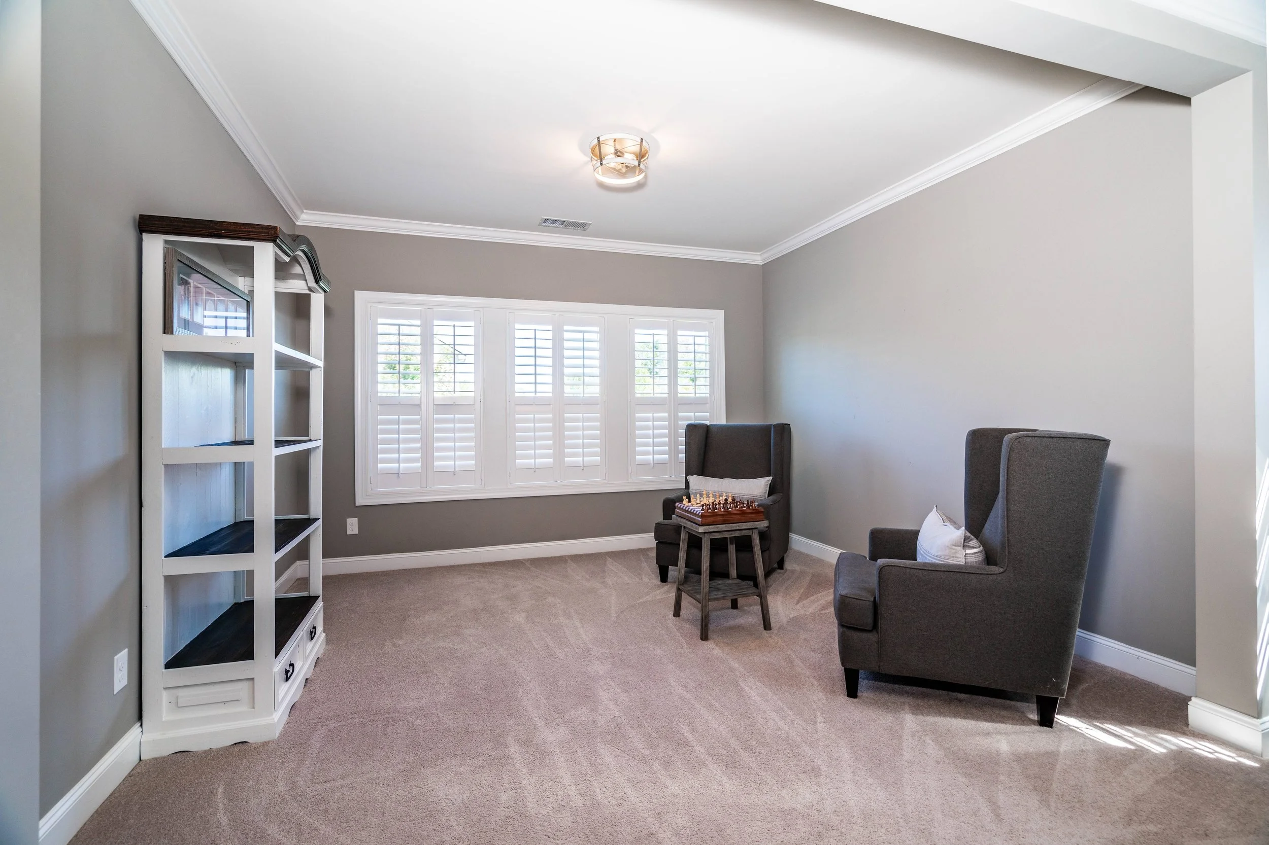 A cozy room with gray walls, a white-framed window with shutters, two plush gray armchairs with cushions, a small table with a chess board, and an empty white bookshelf.