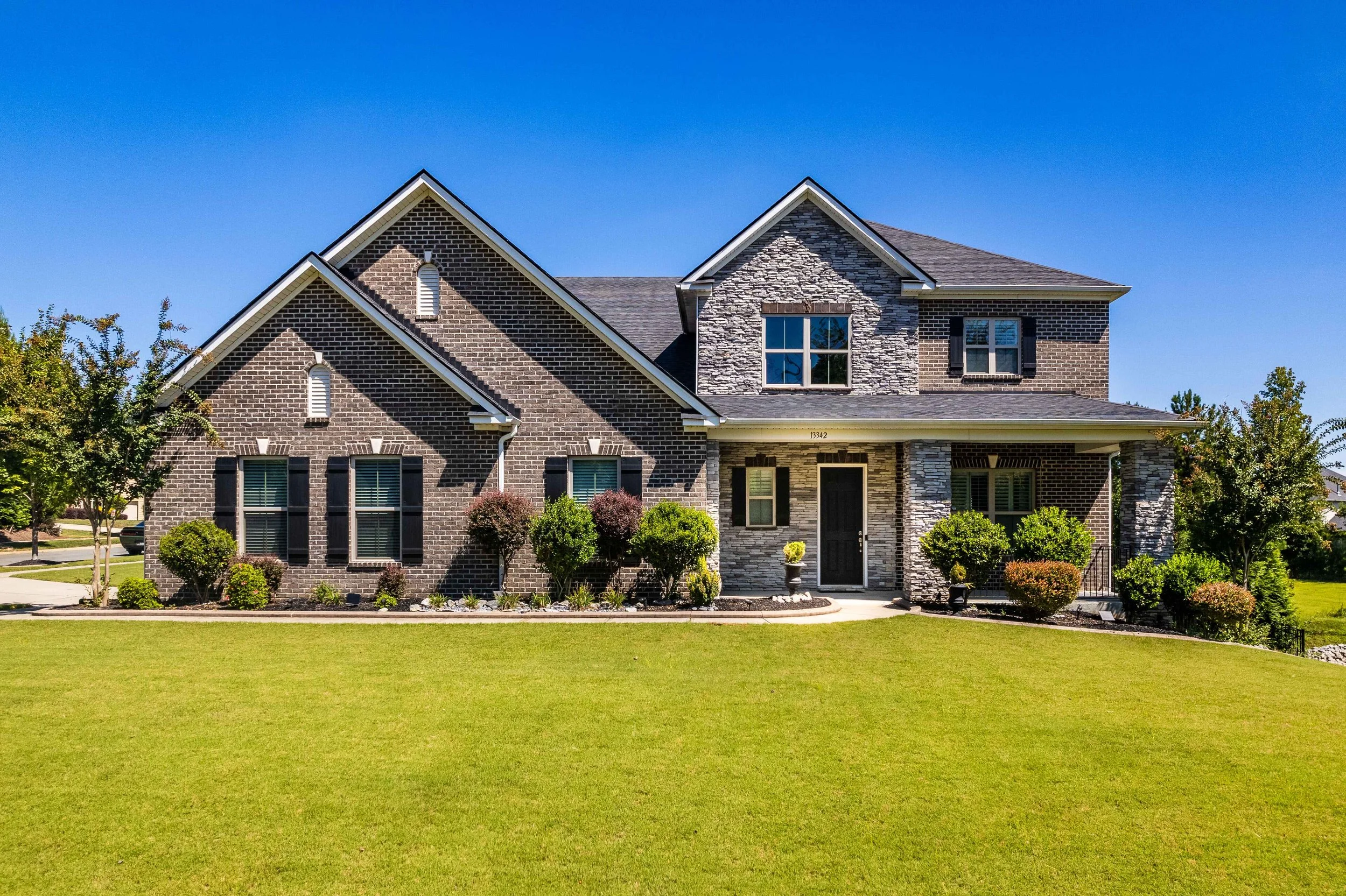 A two-story brick house with a gabled roof, neatly landscaped front yard, and vibrant green lawn under a clear blue sky, conveying a welcoming atmosphere.